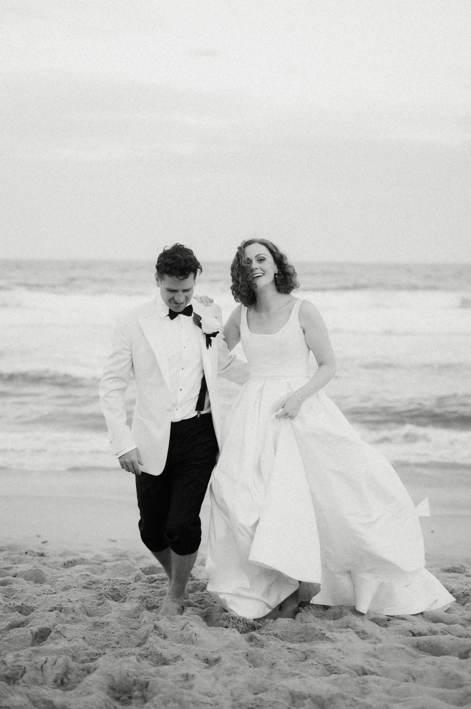 A black and white photo of a bride and groom walking along the beach, smiling and holding hands, with the ocean in the background.