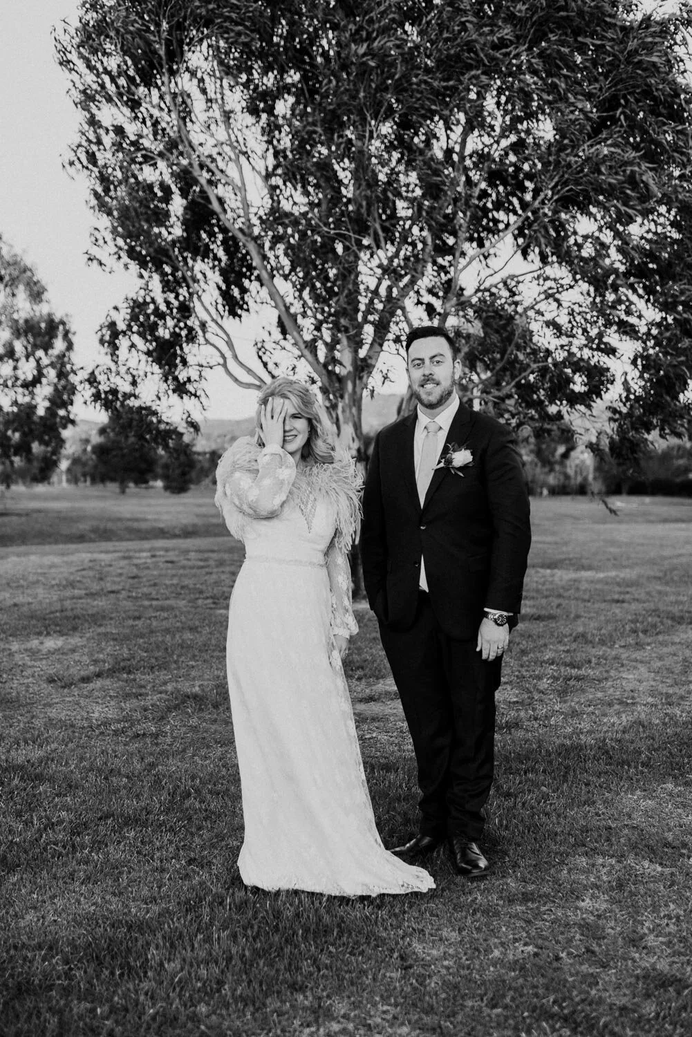 Black and white photo of a wedding couple, a woman in a long dress with feathered accents and a man in a suit with a boutonniere, standing outdoors near a tree, with a grassy field in the background.
