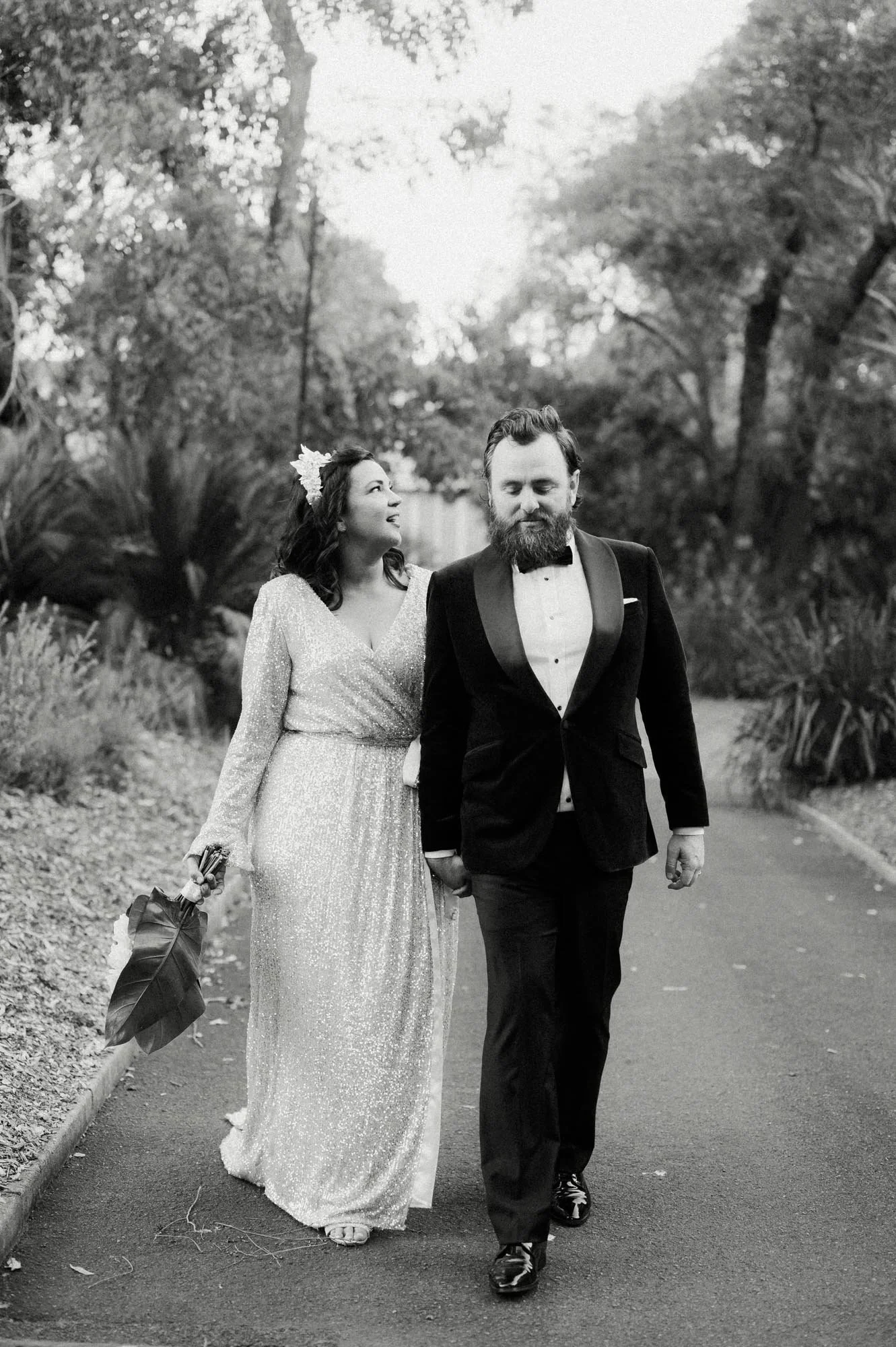 A black-and-white photo of a couple walking arm-in-arm on a park path during daylight. The woman wears a long, sparkly dress with long sleeves and holds a bouquet, and is looking at the man with a smile. The man wears a tuxedo with a bowtie and is lo