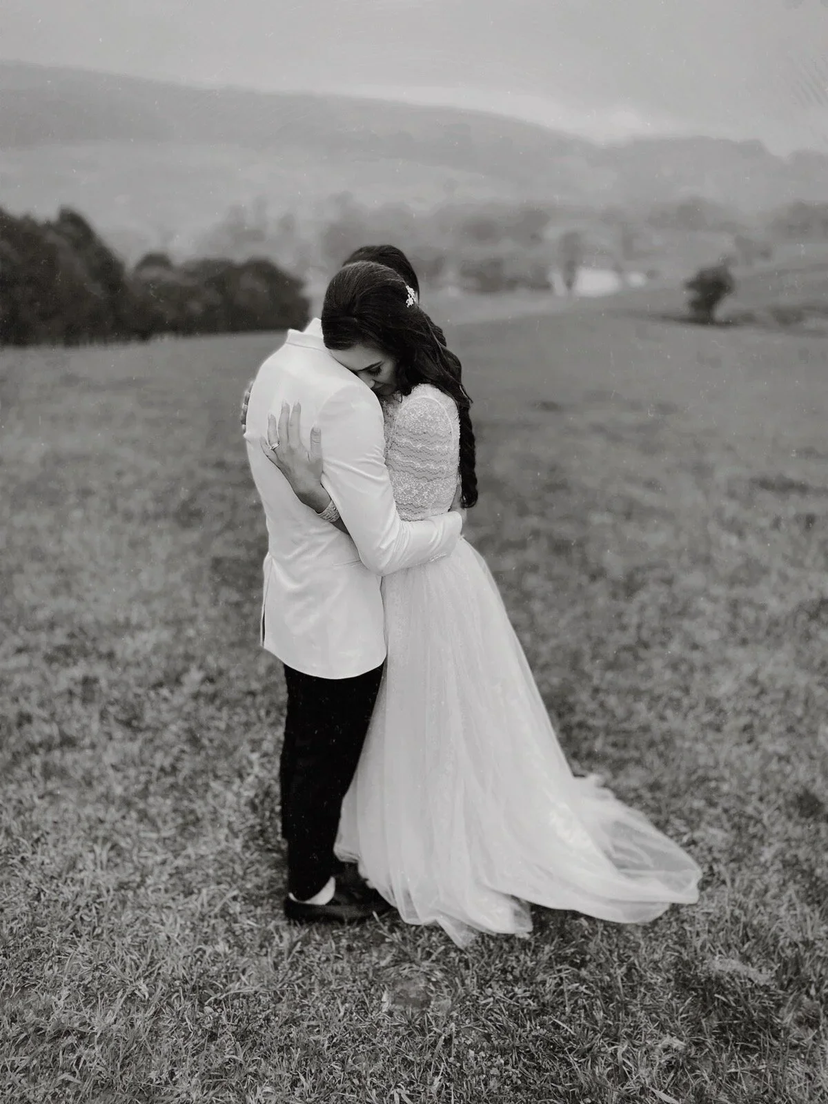 A couple embracing outdoors, with the woman in a wedding dress and the man in a suit, on a grassy field with a blurred landscape in the background.