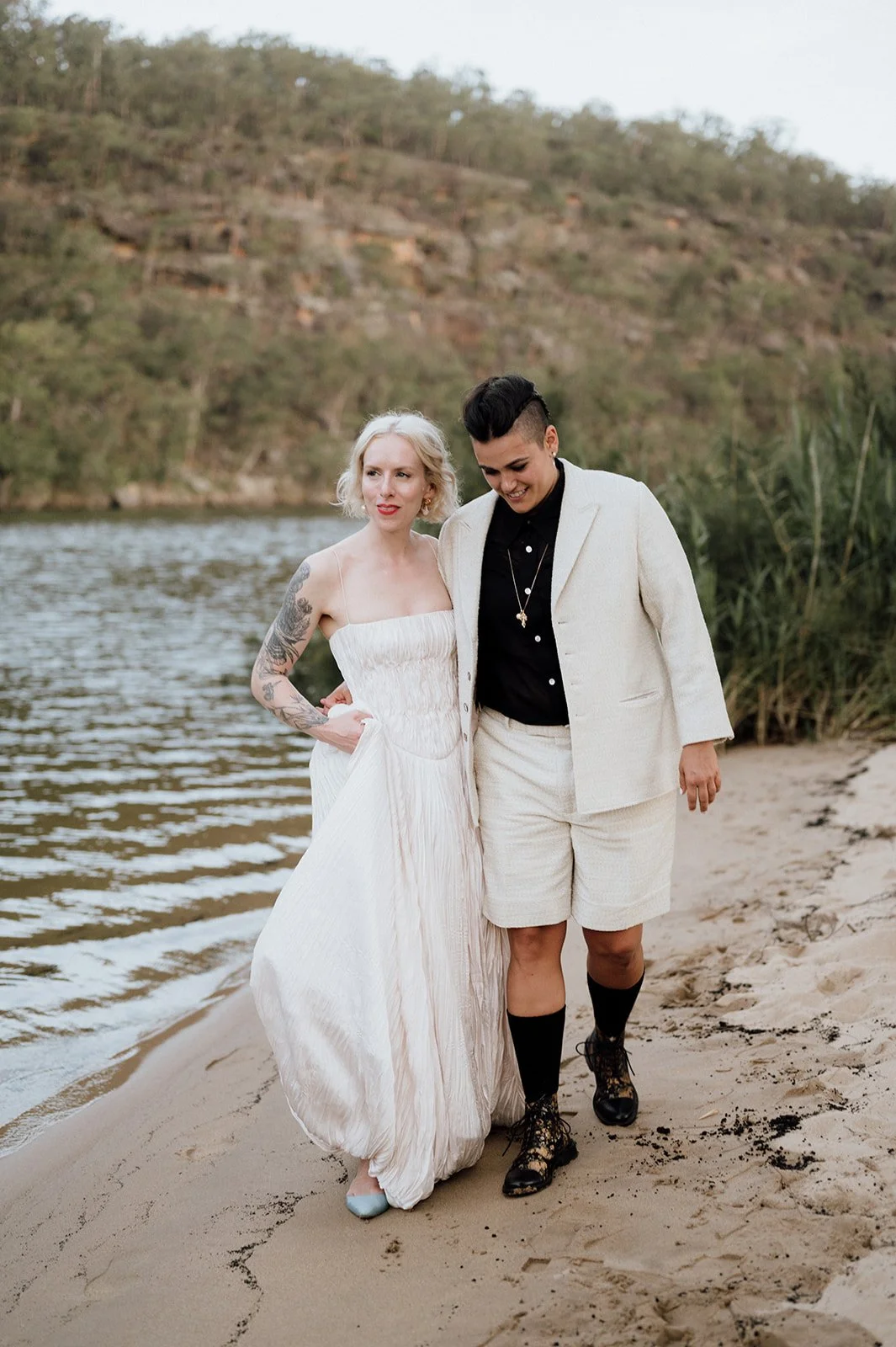 Two same sex brides walking along a riverbank with hills and trees in the background, one in a strapless white dress and the other in a cream-colored suit with black shirt and boots.