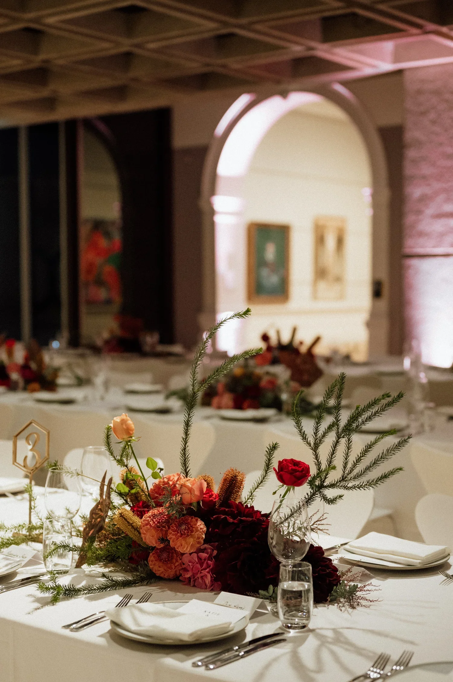 Elegant dining table decorated with a floral centerpiece consisting of red, pink, and peach flowers, surrounded by greenery, set with white plates, silverware, wine glasses, and water glasses. The background shows a softly lit room with artwork on th
