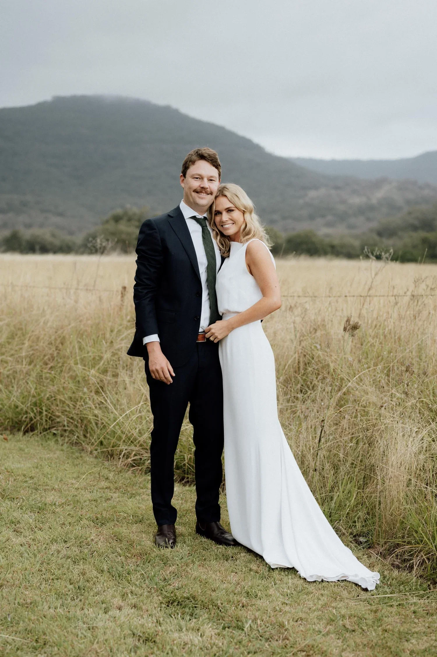 A couple dressed in wedding attire standing outdoors in a grassy field with mountains in the background. The man is wearing a black suit, white shirt, and dark tie, while the woman is in a white wedding dress. They are smiling and posing for the phot