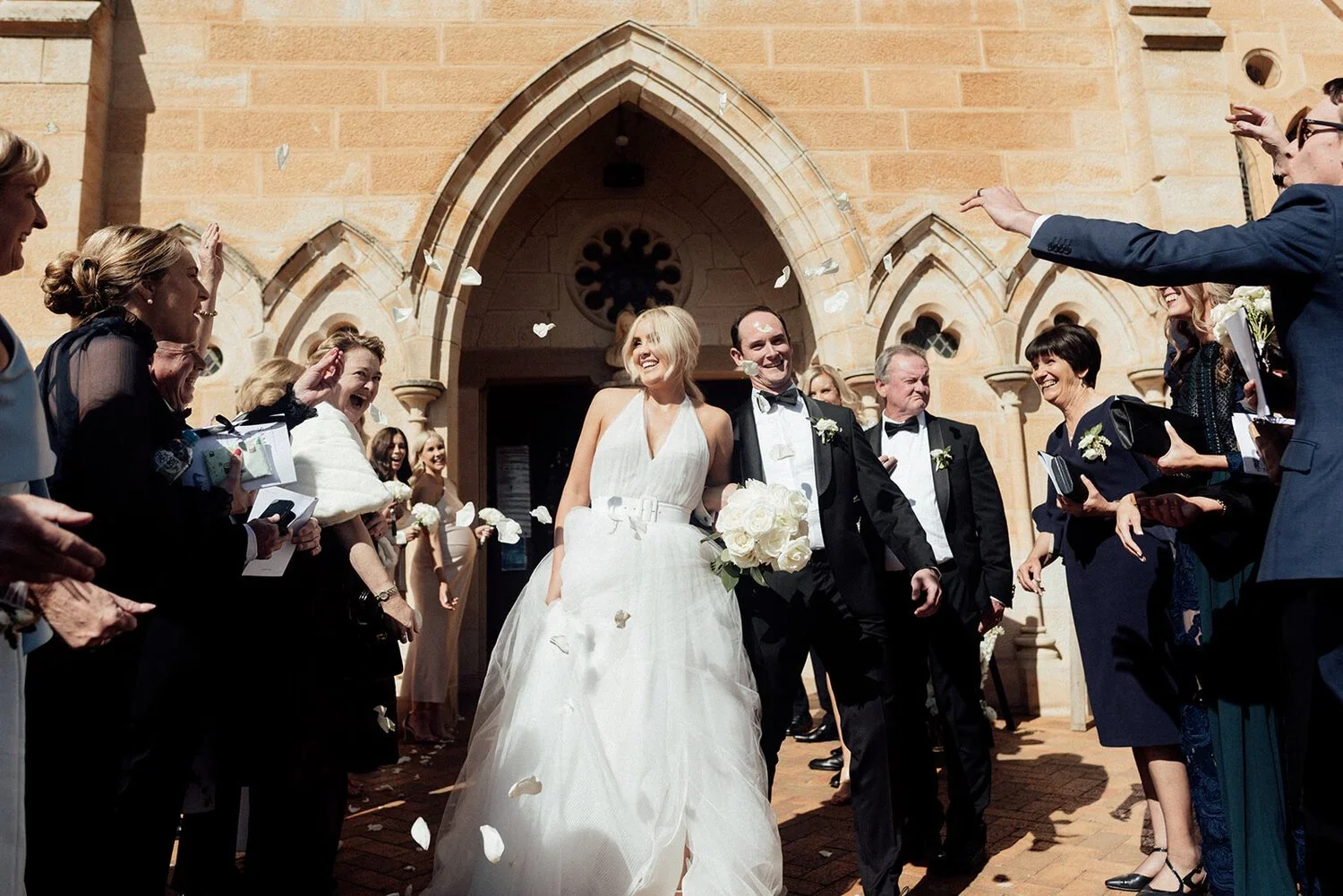 A newlywed couple is leaving a church, smiling and walking together, surrounded by friends and family throwing confetti, in what appears to be a wedding celebration.