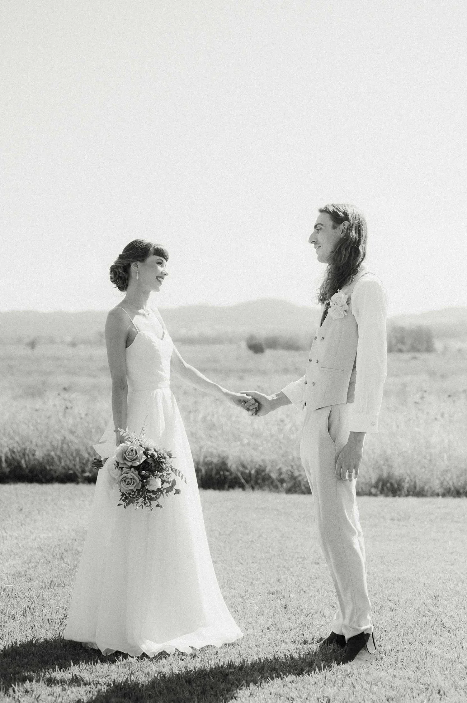 A bride and groom holding hands in a field, facing each other, smiling, with a bouquet in the bride's hand, during a wedding ceremony.