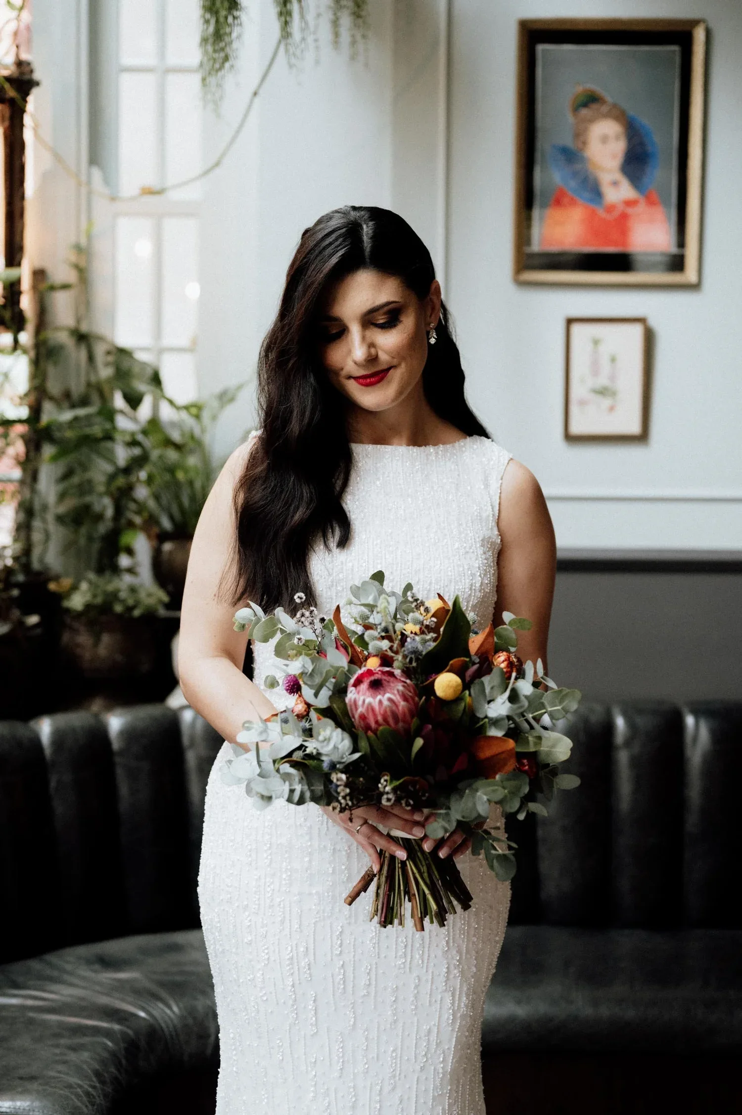 A woman in a white wedding dress holding a bouquet of flowers, standing indoors with artwork and plants in the background.