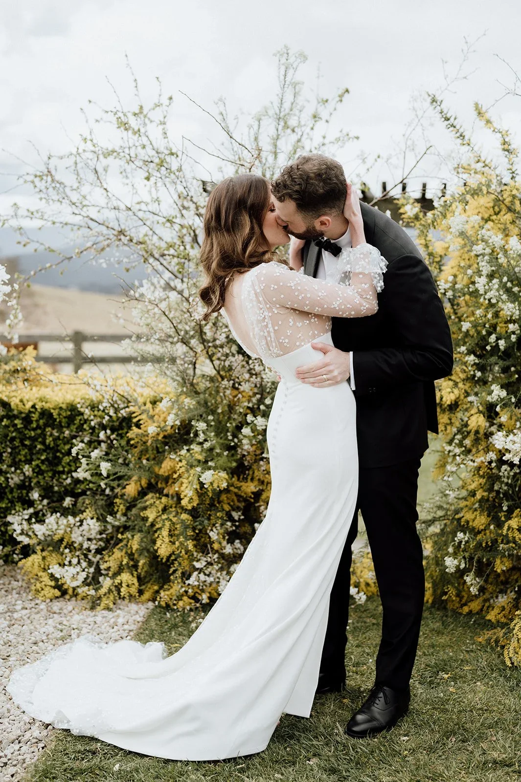 A bride and groom sharing a kiss outdoors, with the bride wearing a white wedding gown with sheer, floral-embellished sleeves and the groom in a black tuxedo, surrounded by blooming yellow and white flowers - Seclusions Blue Mountains photography.