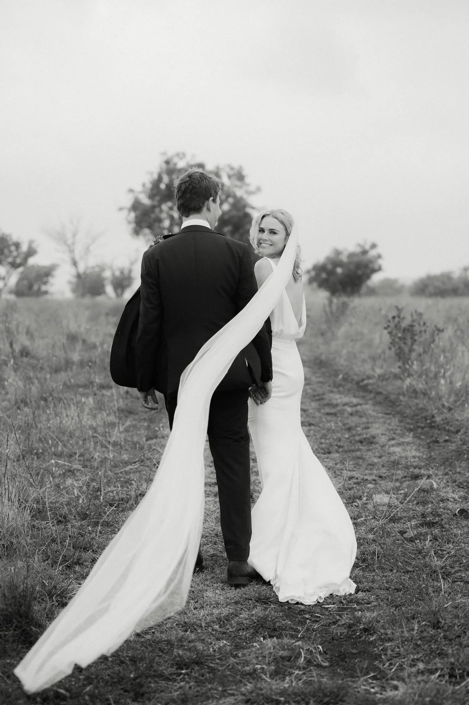 Black and white photo of a bride and groom holding hands, walking in an open field, with trees in the background, the bride smiling at the camera, the groom facing away.