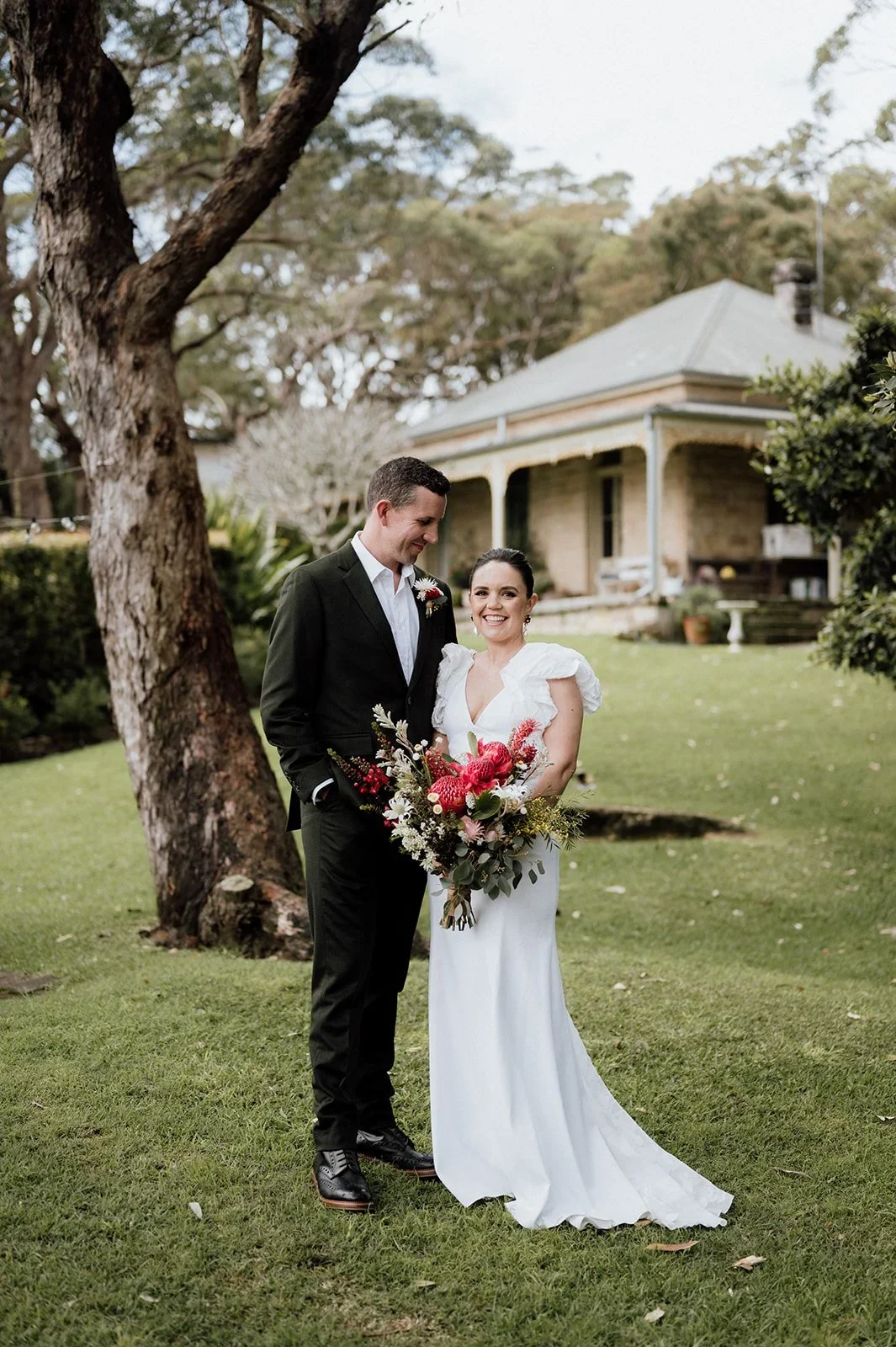 A bride in a white dress holding a large bouquet, smiling, standing next to a groom in a black suit. They are outdoors on a grassy area near a large tree, with a house in the background.