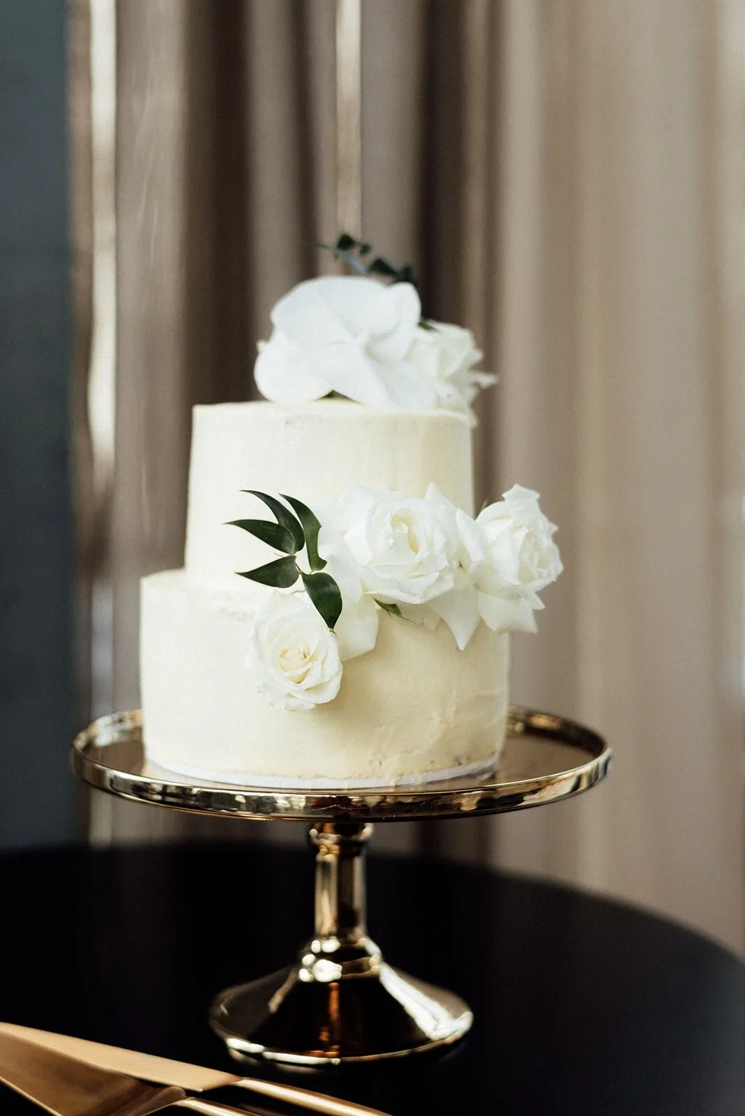 A white, two-tiered wedding cake decorated with white roses and greenery on a silver cake stand - inner city wedding photographer in Sydney.