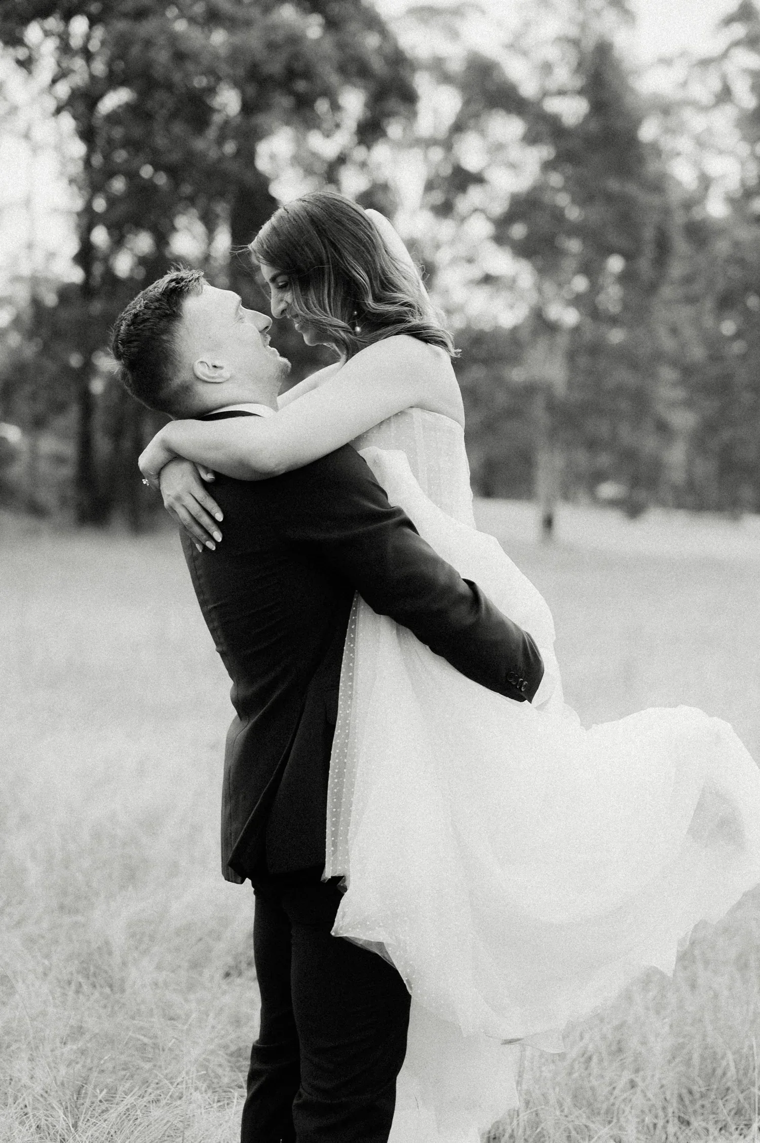 A black and white photograph of a couple outdoors, with the man lifting the woman in his arms. Both are smiling and looking at each other, with trees in the background.