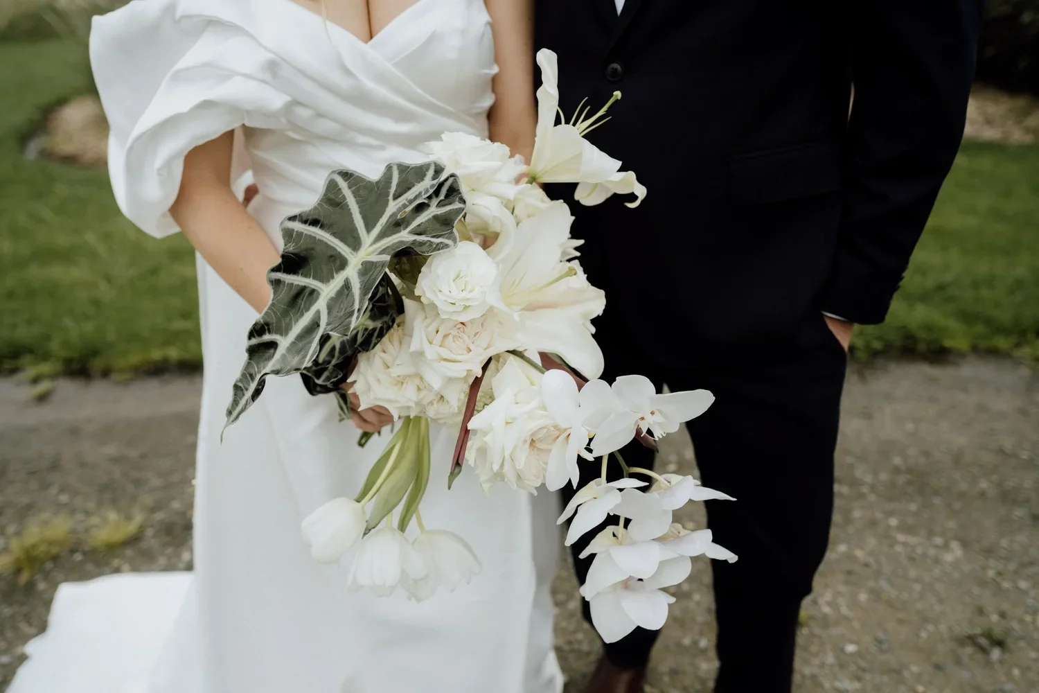Close-up of a bride in a white wedding gown holding a cascading bouquet of white flowers and green foliage, standing next to a groom in a black suit outdoors on a grassy area.