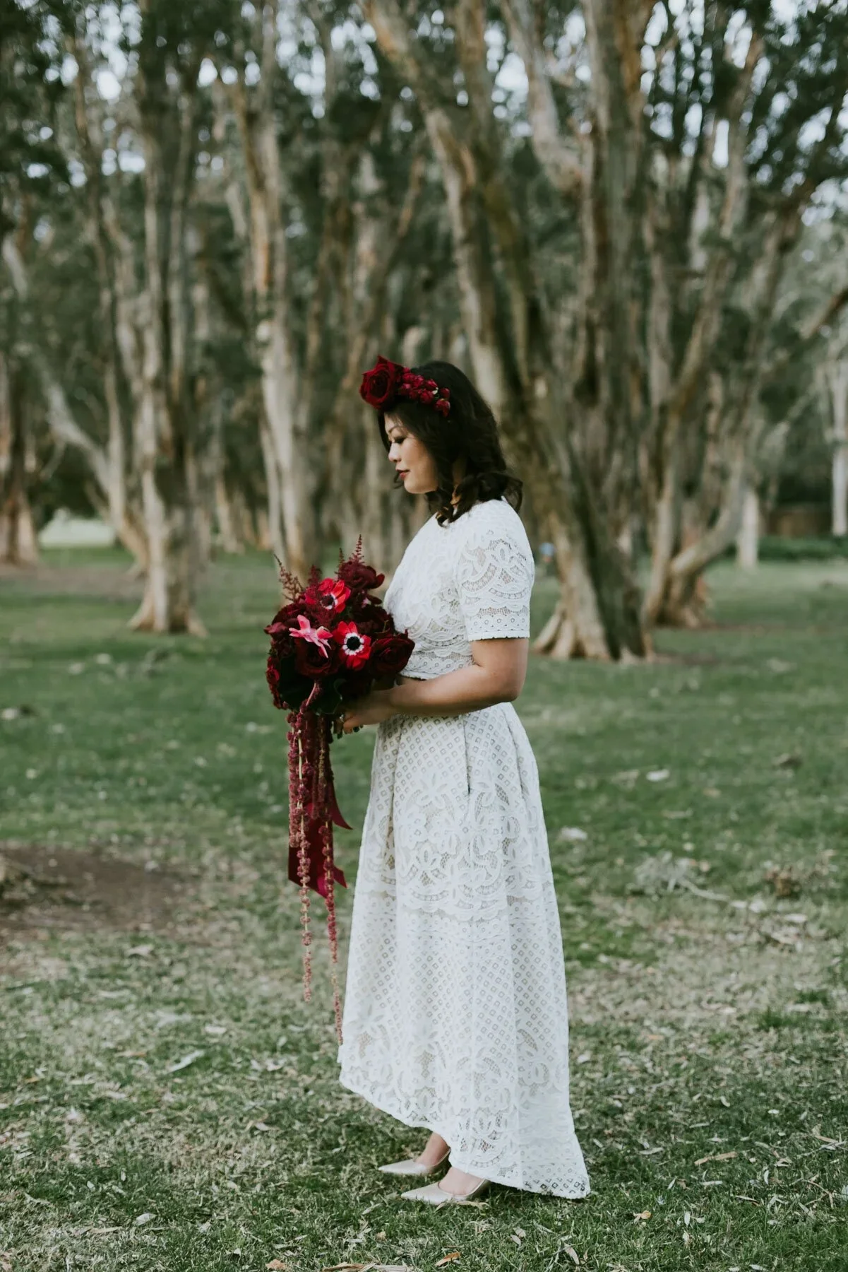Woman in a white lace dress holding a bouquet of red flowers in a park with large trees in the background.