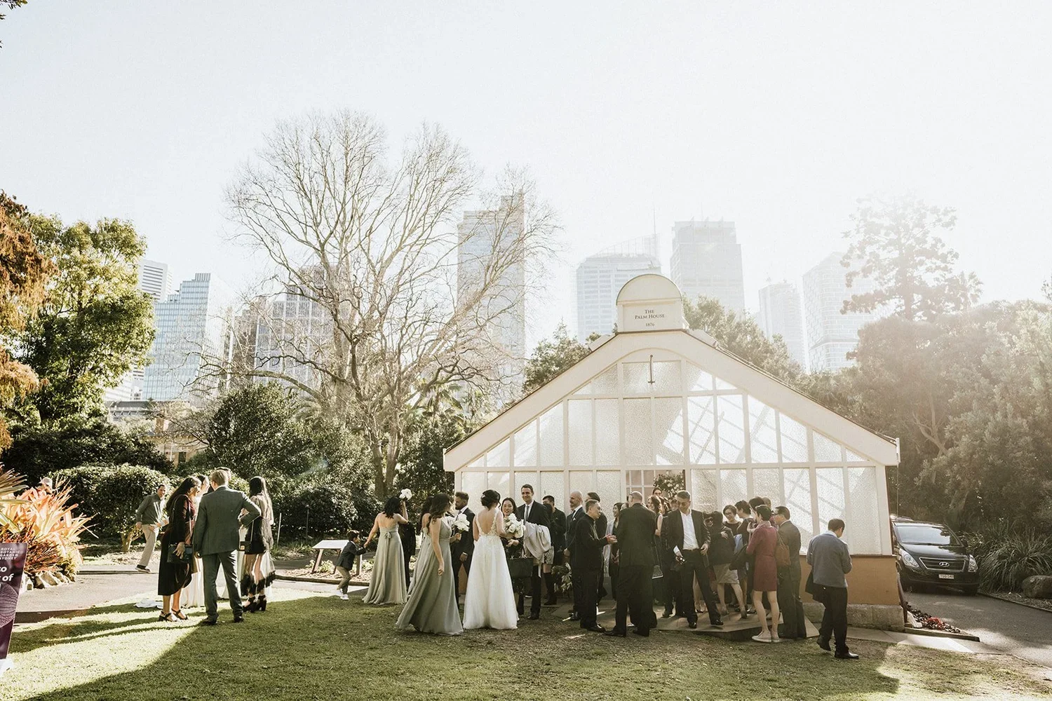 Wedding celebration outside a glass house surrounded by trees and city skyscrapers in the background.