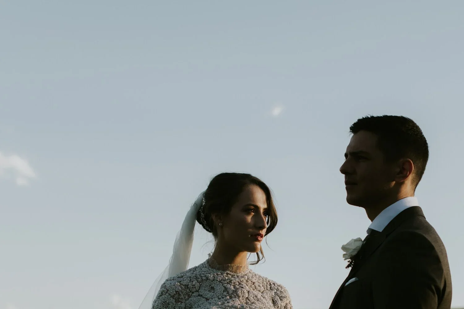 A bride and groom facing each other outdoors during sunset, with clear sky background.