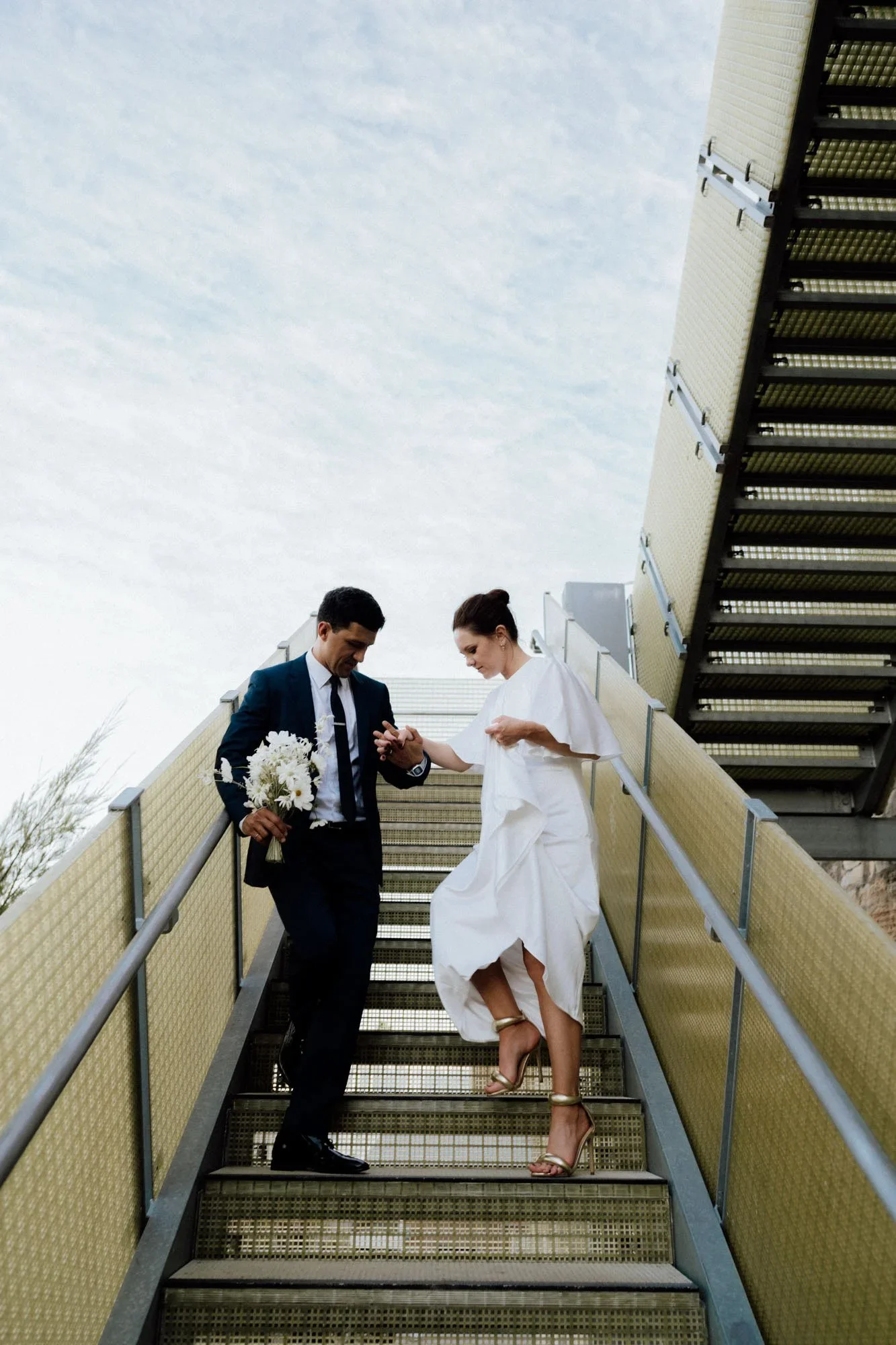 A man in a suit and a woman in a white dress are standing and descending outdoor metal stairs, with the man holding a large bouquet of white flowers. The woman is lifting her dress slightly, revealing high-heeled sandals. They appear to be engaged in