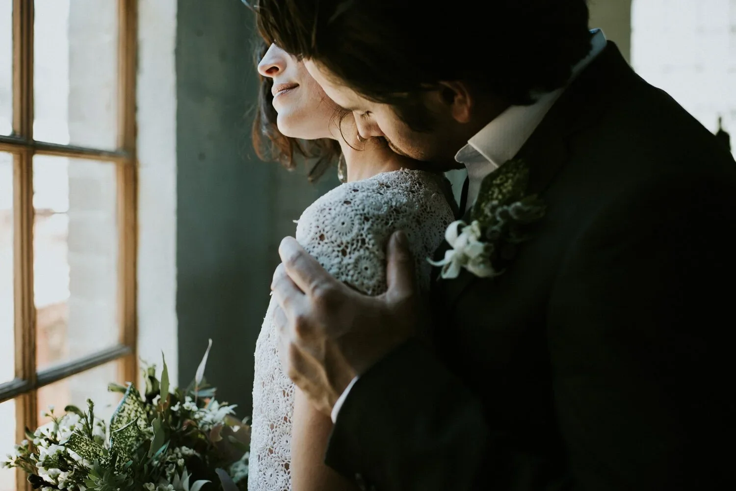 A couple embracing on their wedding day indoors near a window, with sunlight coming in, the man is kissing the woman's shoulder, she is wearing a lacy dress and he is in a suit with a boutonniere.