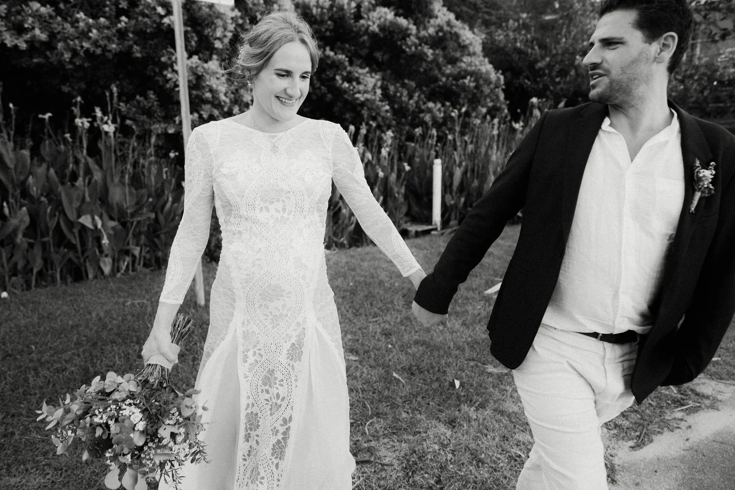 A smiling woman in a lace wedding dress holding a bouquet, holding hands with a man in a black blazer and white shirt, walking outdoors on grass with trees in the background.