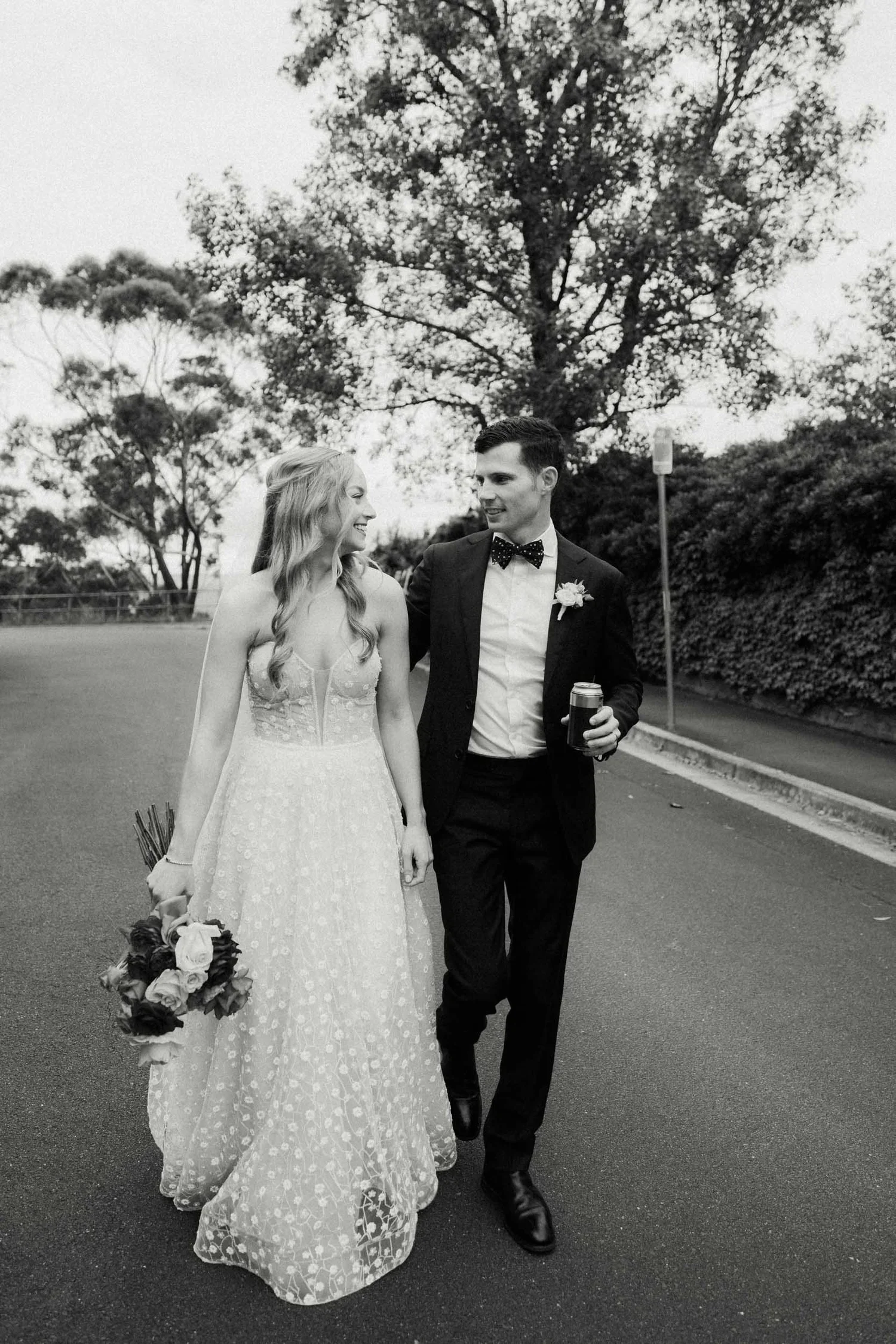 A bride and groom walking together outdoors. The bride is holding a bouquet of flowers and wearing a lace wedding dress. The groom is dressed in a suit with a bow tie and is holding a drink. They are smiling at each other and walking on a street with