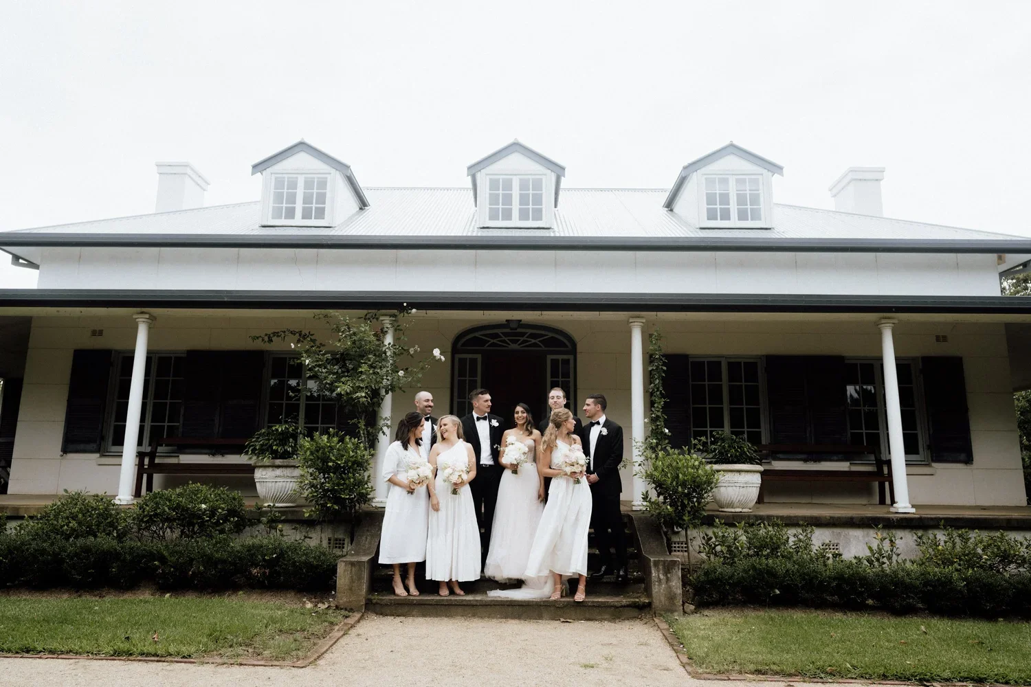 A group of wedding guests standing on the steps of a large, white, two-story house with dormer windows and a porch. The group includes women in white dresses holding bouquets and men in tuxedos, smiling and talking.