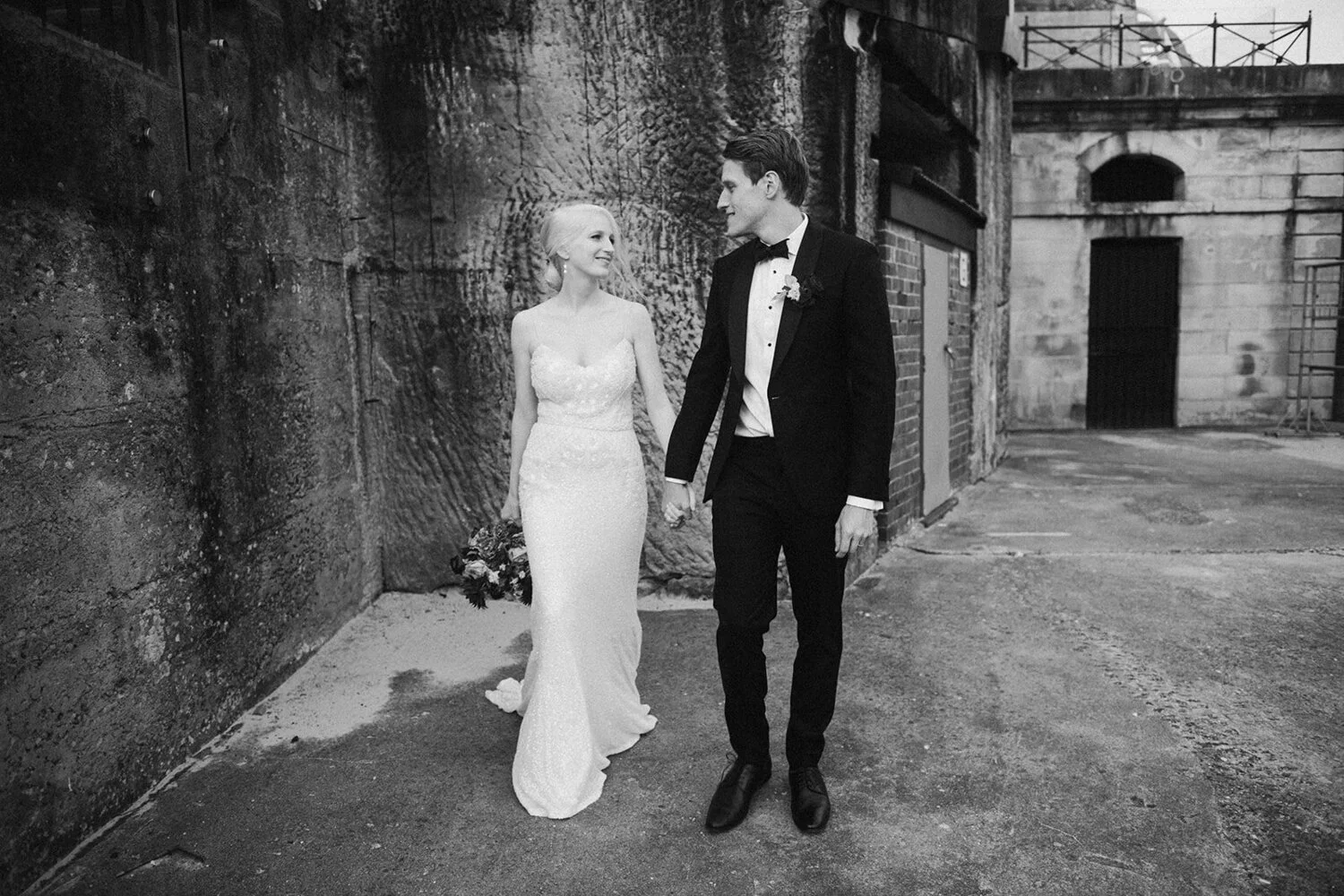 A bride and groom walking hand in hand in an urban setting, with the bride wearing a white wedding dress and the groom in a black tuxedo, both smiling at each other.
