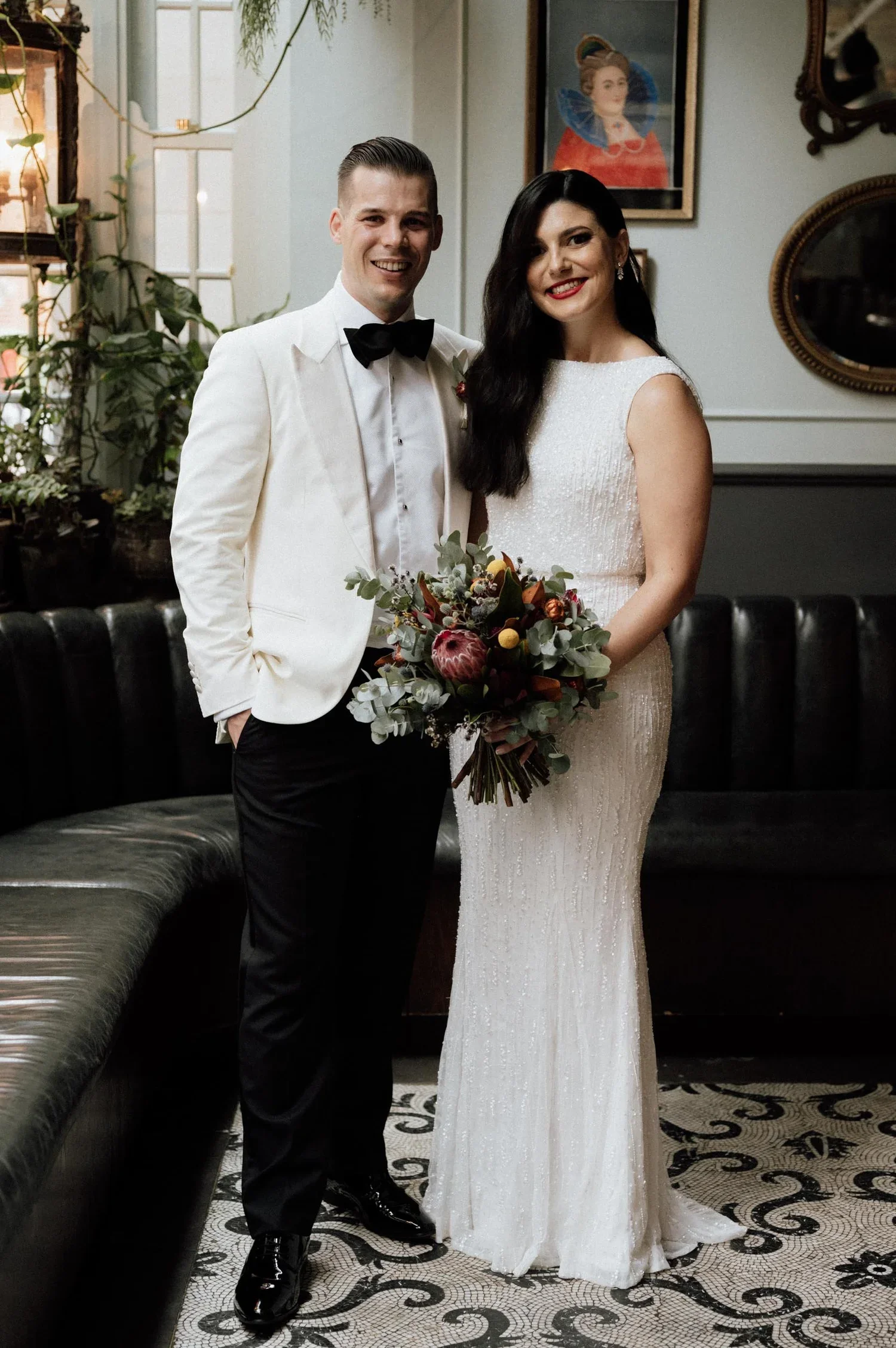 A groom and bride standing together indoors, smiling at the camera. The groom is wearing a white tuxedo with a black bow tie, and the bride is in a white, sleeveless, beaded wedding gown holding a bouquet of flowers. Behind them are framed pictures a