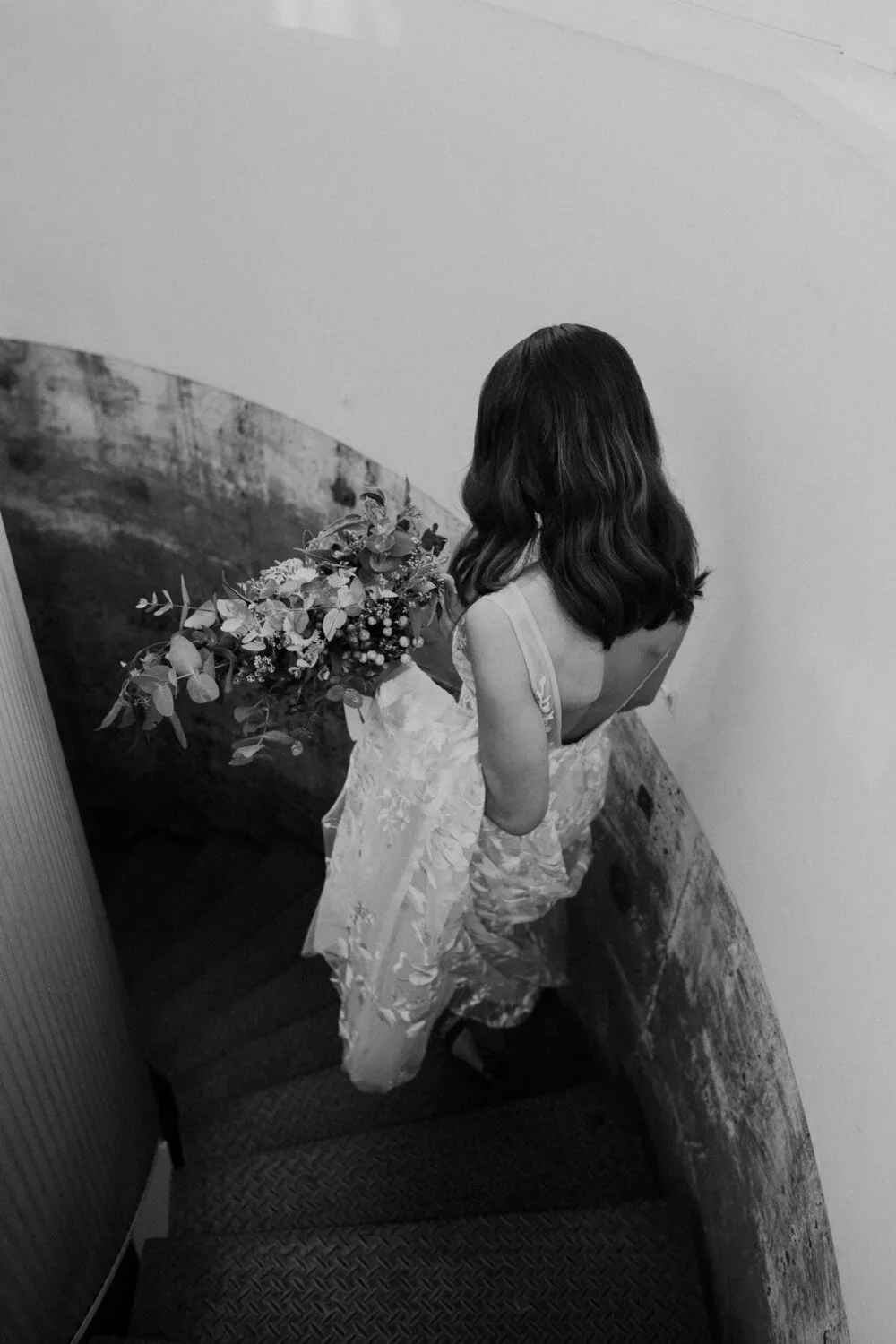 A woman with wavy hair, wearing a floral dress, holding a bouquet of flowers, standing on a staircase with a curved brick wall.