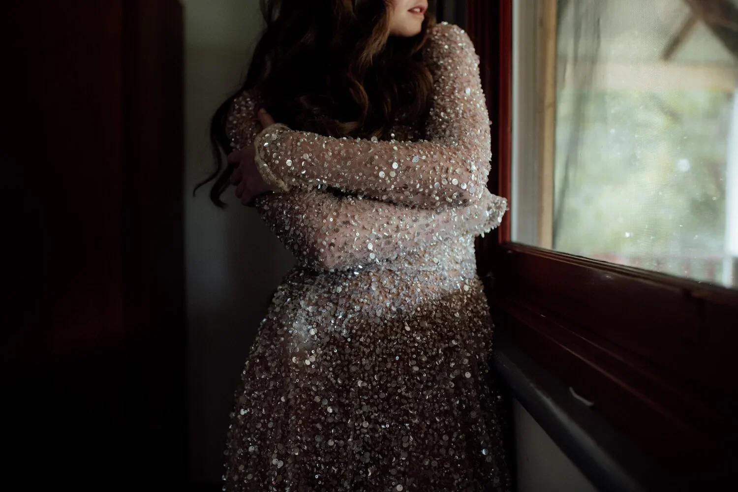 Woman wearing a long sleeve dress covered in sequins, standing by a window with arms crossed, looking outside.