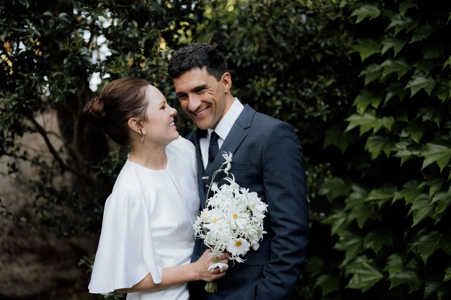 A bride and groom smiling at each other outdoors, the bride holding a bouquet of white daisies and white orchids, surrounded by greenery.