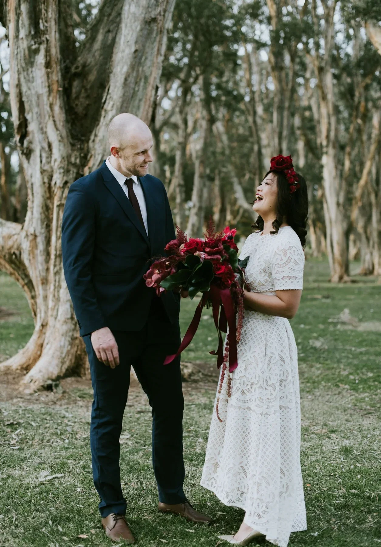 A couple stands outdoors in a wooded area, with the woman wearing a white lace dress and flower crown, and the man in a dark suit. The woman is holding a bouquet of red flowers and is smiling at the man, who is looking at her. The scene suggests a we
