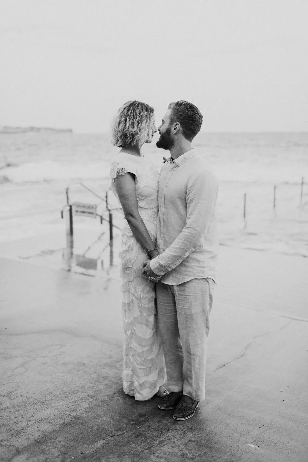 A couple standing close together on the beach, face-to-face, holding hands, with the ocean in the background.
