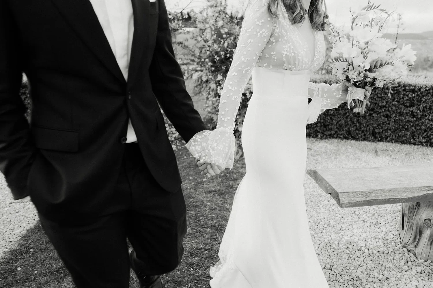 A couple dressed in wedding attire holding hands, with the woman holding a bouquet, walking outdoors.