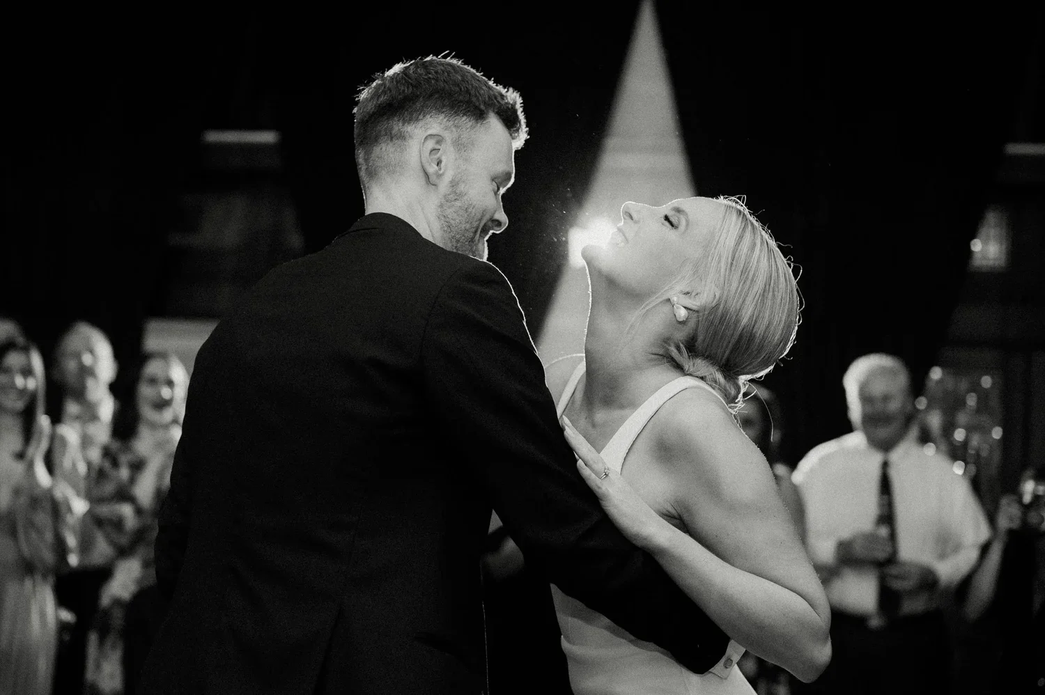 A couple dancing closely at a wedding reception with friends watching in the background, black and white photo.