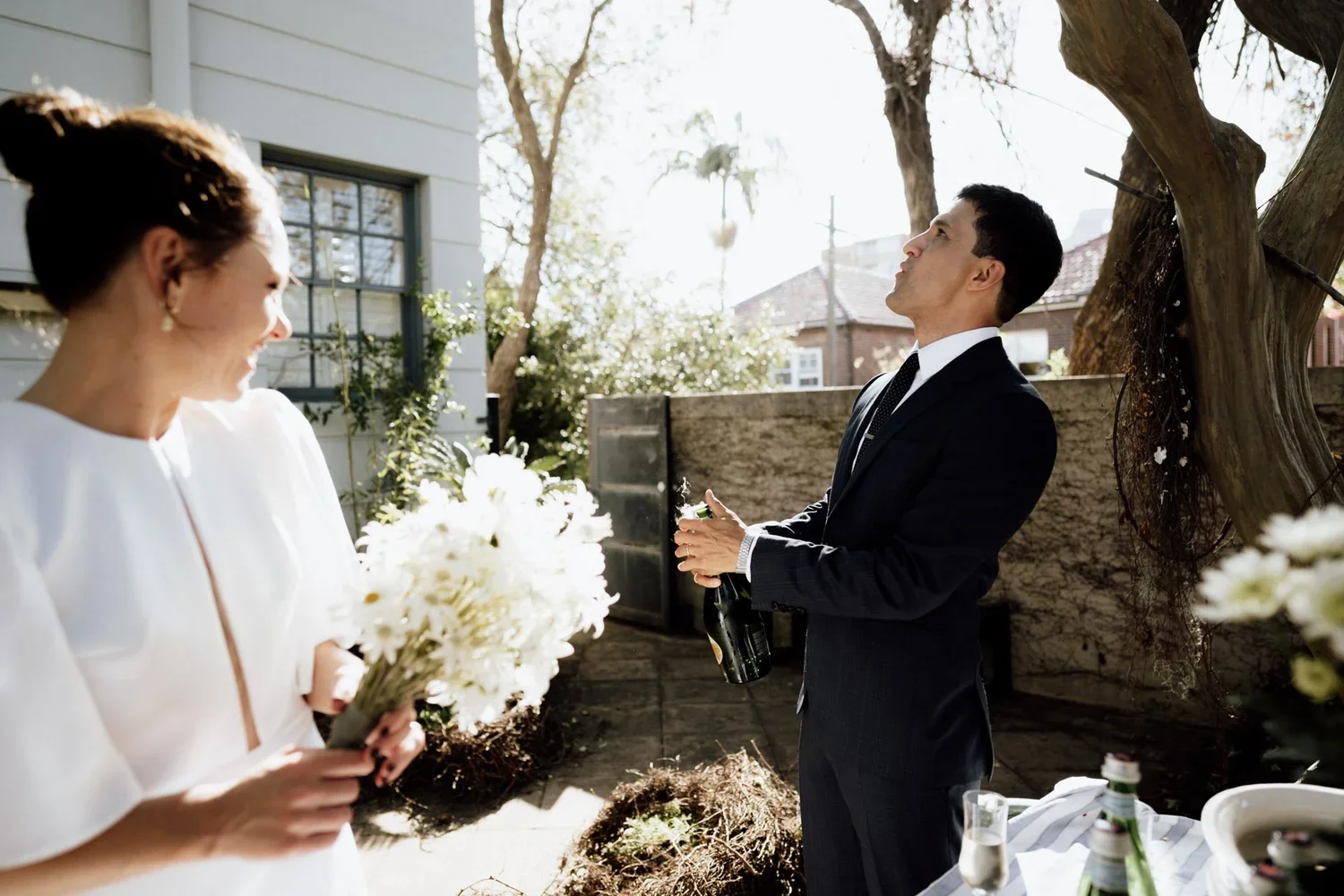 A woman holding a bouquet of white flowers and a man in a suit holding a bottle of champagne, standing outside in a backyard with trees and a wooden fence, during daytime.