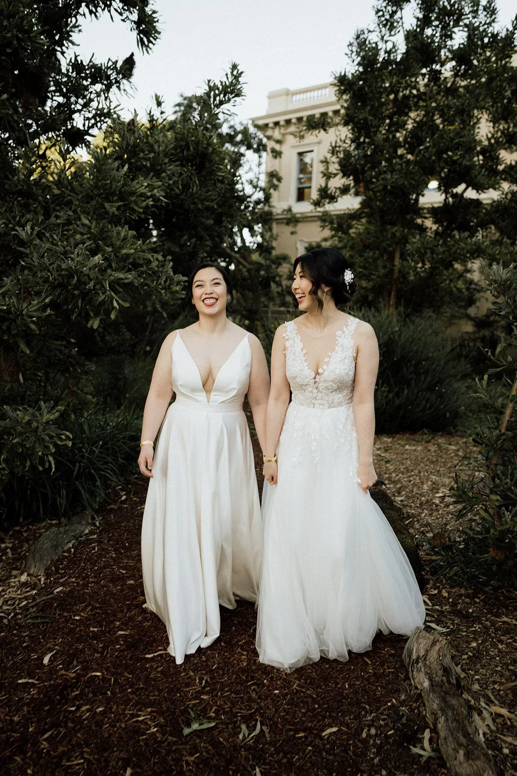 Two women in wedding dresses walking hand-in-hand outdoors, smiling and looking at each other, surrounded by greenery and a building in the background.