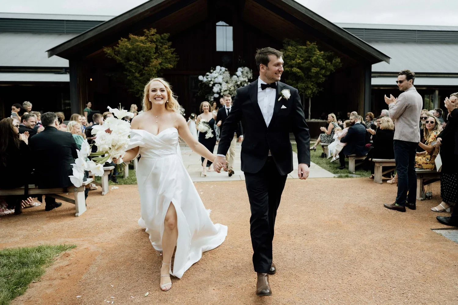 A newlywed couple walking hand in hand at their outdoor wedding ceremony. The bride is holding a bouquet of white flowers and wearing a strapless white wedding gown, while the groom is in a black tuxedo with a bow tie. Guests are seated and clapping 