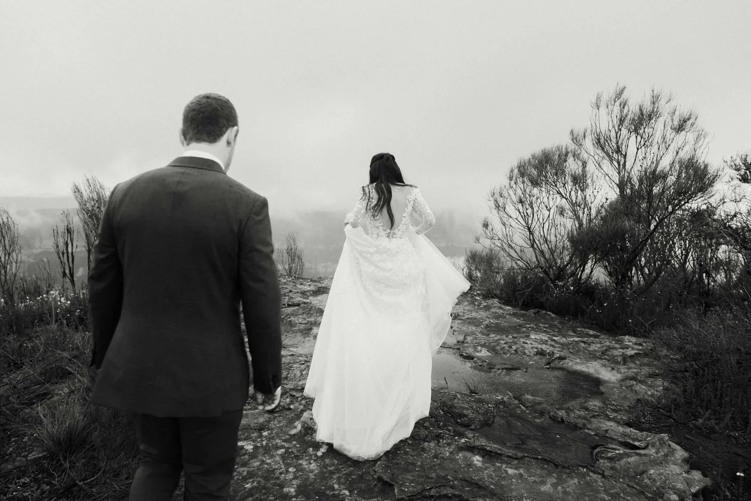 A black-and-white photo of a bride in a lace wedding dress walking away on a rocky path, with a man in a suit following behind, on a foggy outdoor landscape with sparse trees.