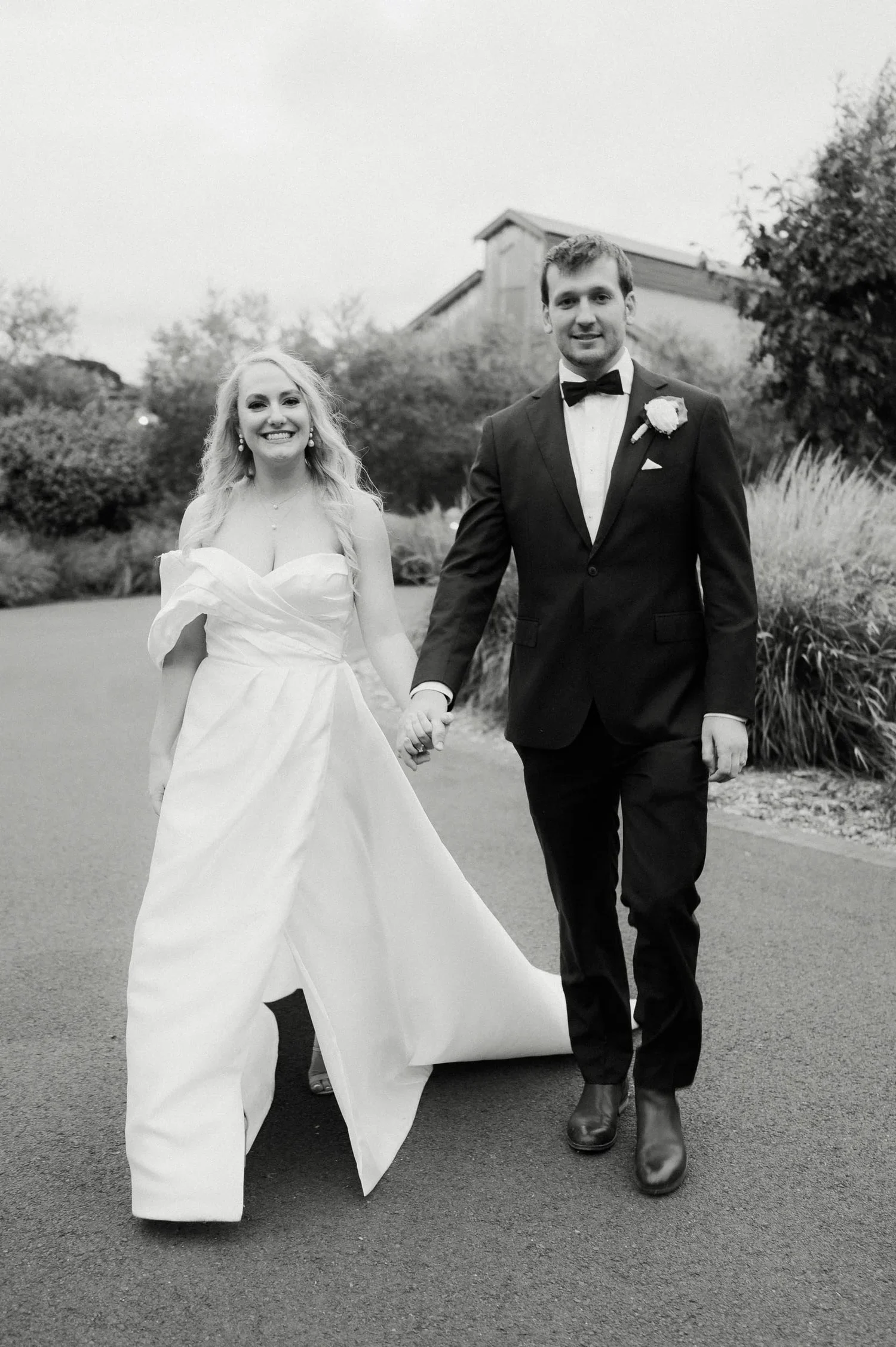 A black and white photo of a smiling bride and groom walking hand in hand outdoors. The bride is wearing a strapless wedding gown with a large bow detail and a necklace, while the groom is dressed in a black tuxedo with a bow tie and a flower boutonn