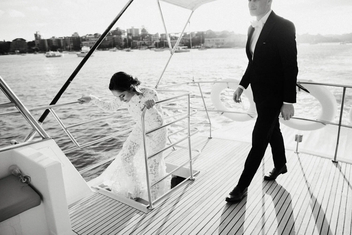 A woman in a lace wedding dress is boarding a yacht, with a man in a suit and bow tie walking nearby on the deck. The scene is set on a body of water with boats and buildings in the background.