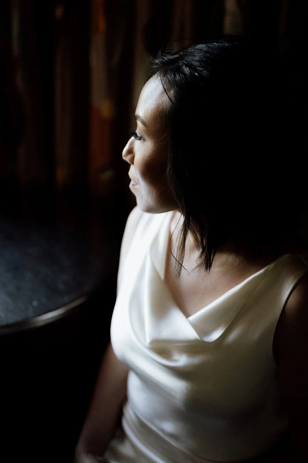 A woman with black hair and light skin sitting near a window, wearing a cream-colored dress, with her face turned sideways in profile, illuminated by soft natural light.