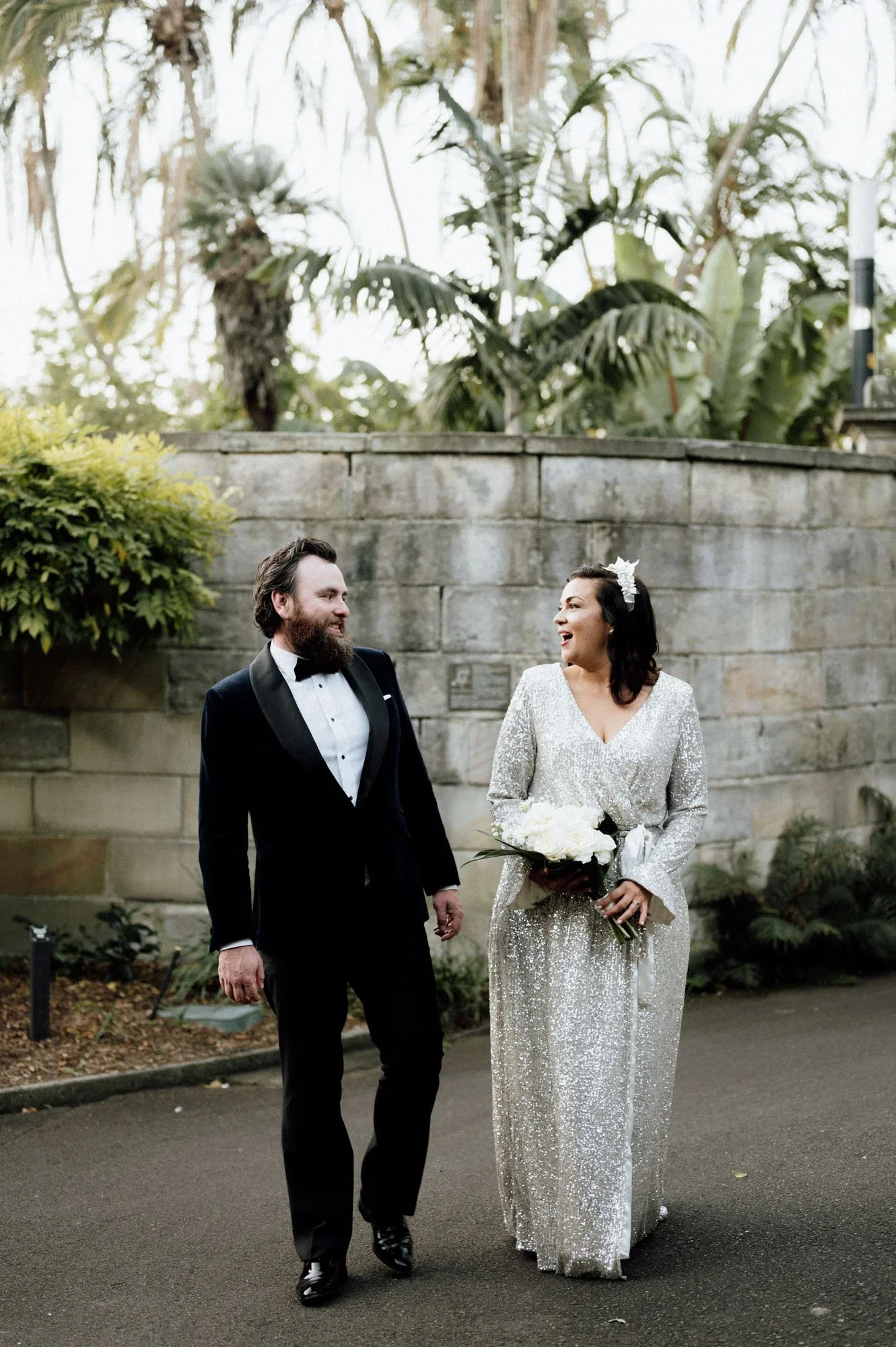 A newlywed couple walking outside in front of a stone wall, holding hands and smiling at each other. The groom is wearing a black tuxedo with a white shirt and bow tie. The bride is wearing a sparkling silver wedding gown, holding a bouquet of white 
