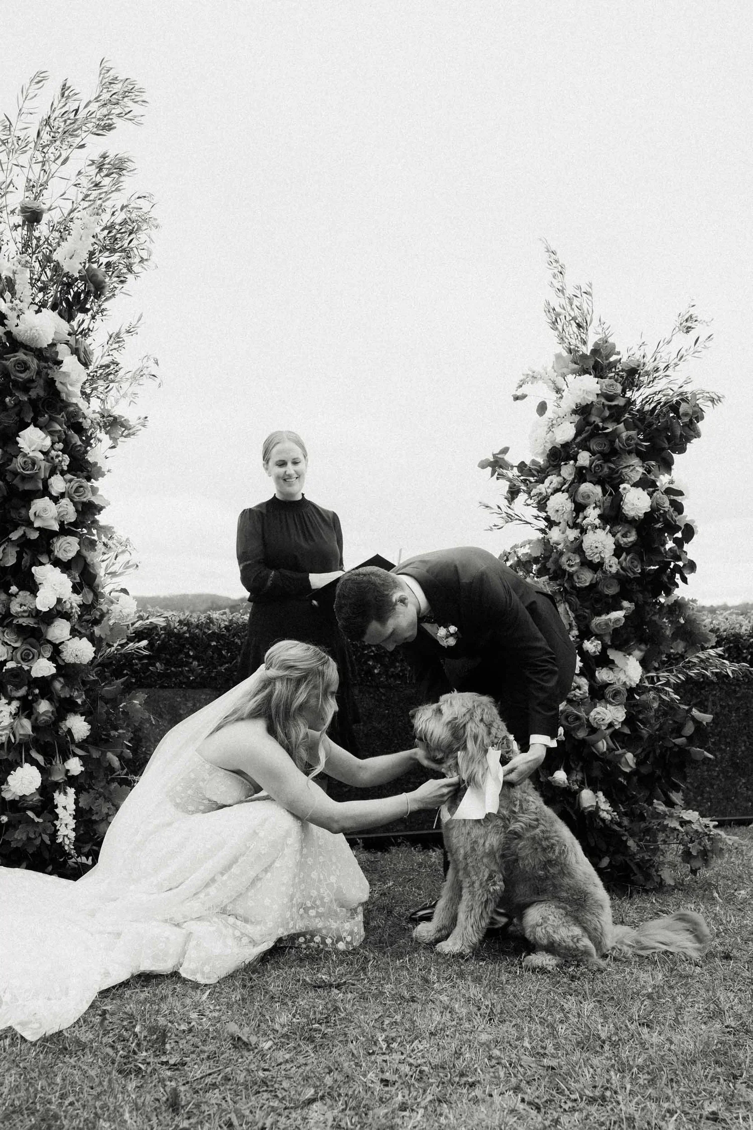 A wedding ceremony outdoors with floral arches, a bride in a white dress, a groom in a suit, and a woman holding a dog, with a woman in a black dress standing nearby.