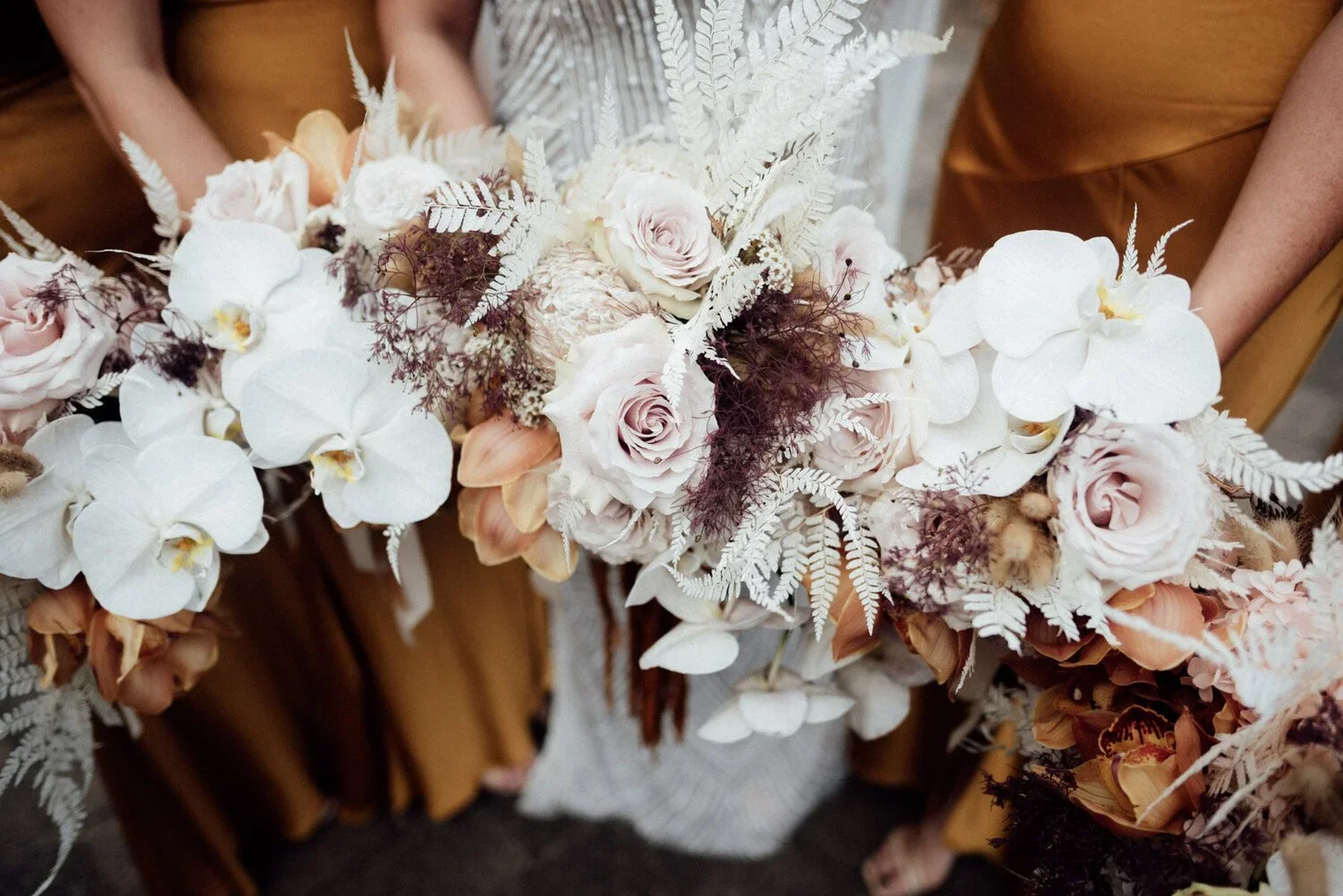 A bouquet of white orchids, blush roses, ferns, and dark foliage held by people in beige and brown clothing.