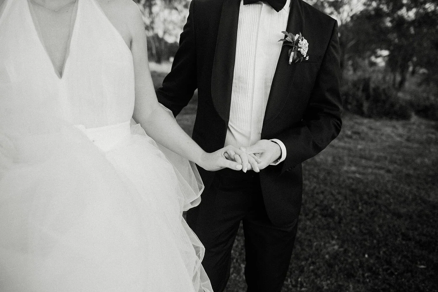 A black and white photograph of a bride and groom holding hands during their wedding, with the focus on their clasped hands. The bride is wearing a sleeveless white wedding dress with a tulle skirt, and the groom is in a black tuxedo with a bow tie a
