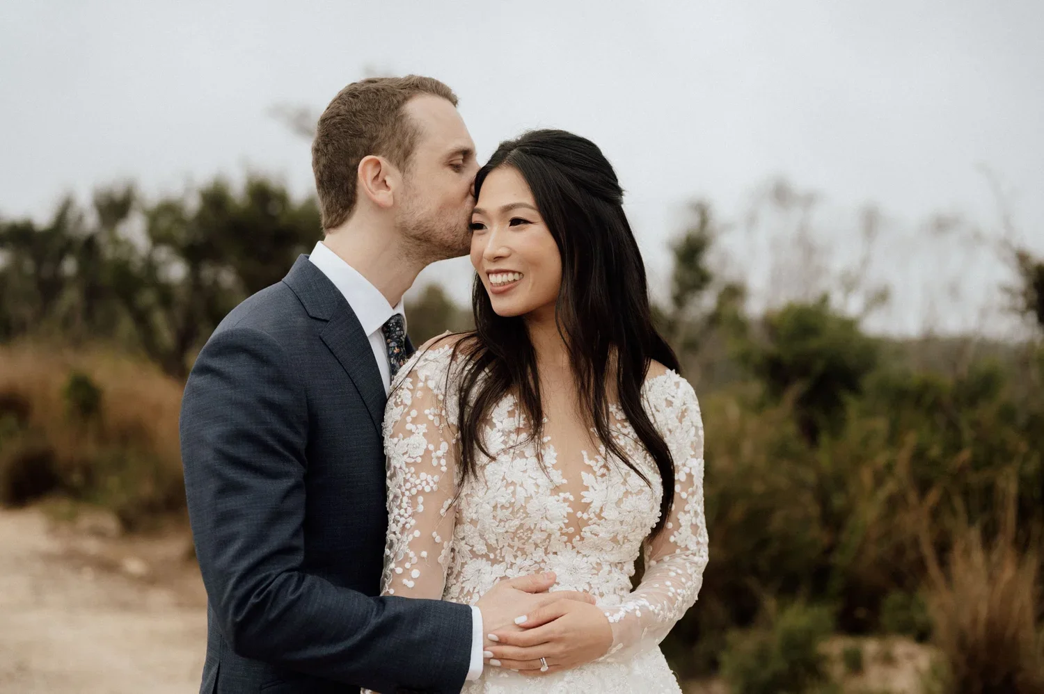 A happy couple on their wedding day; the groom is giving a kiss on the bride's forehead while she smiles, dressed in a wedding gown, outdoors with blurred trees in the background.