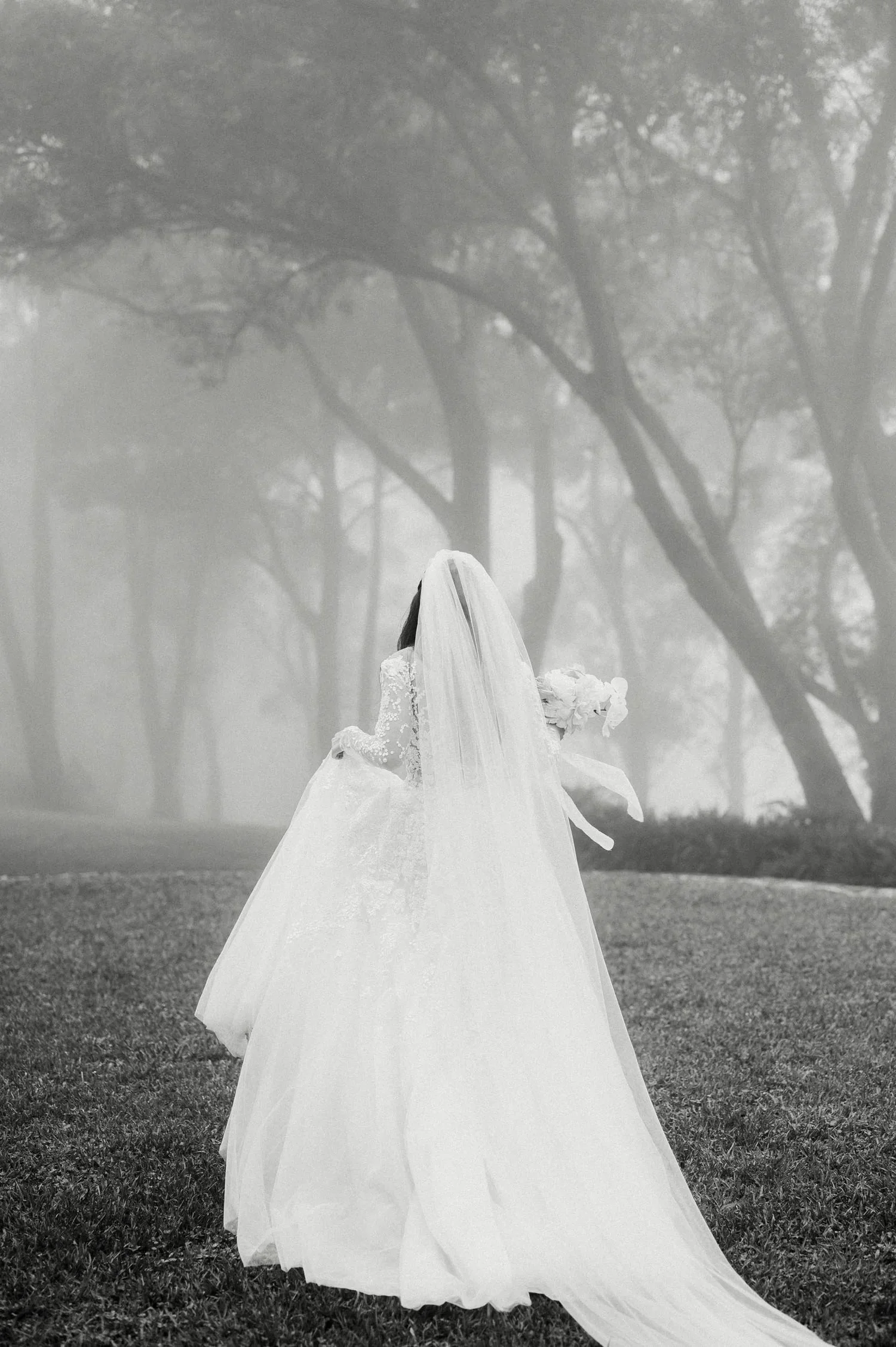 A bride in a wedding dress with a long train, holding a bouquet of flowers, standing outdoors on grass with trees and fog in the background.