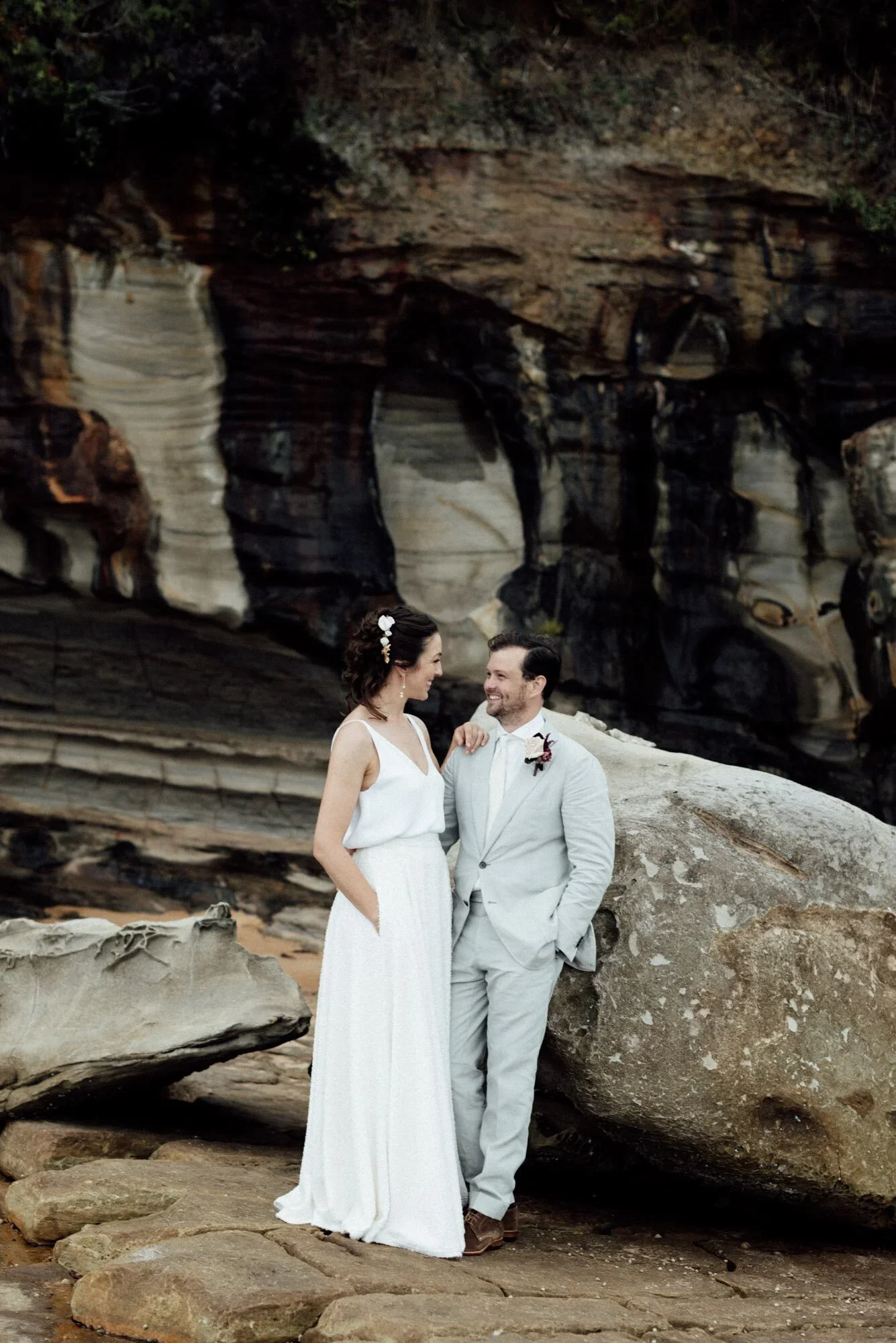 A bride and groom standing on rocks on a beach, smiling and looking at each other, with a large rock formation in the background.