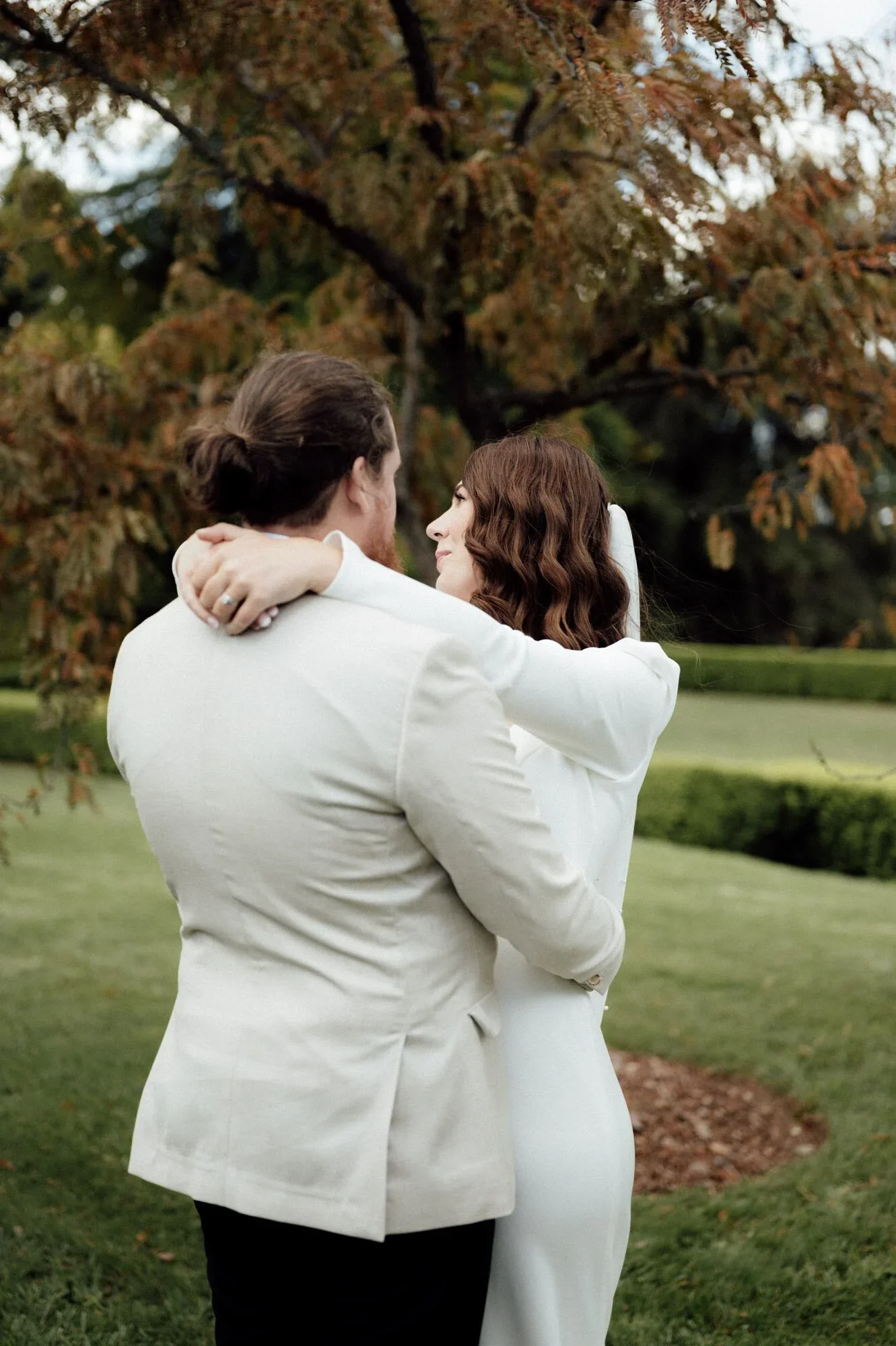 A couple in wedding attire embracing outdoors in front of a tree with autumn leaves.