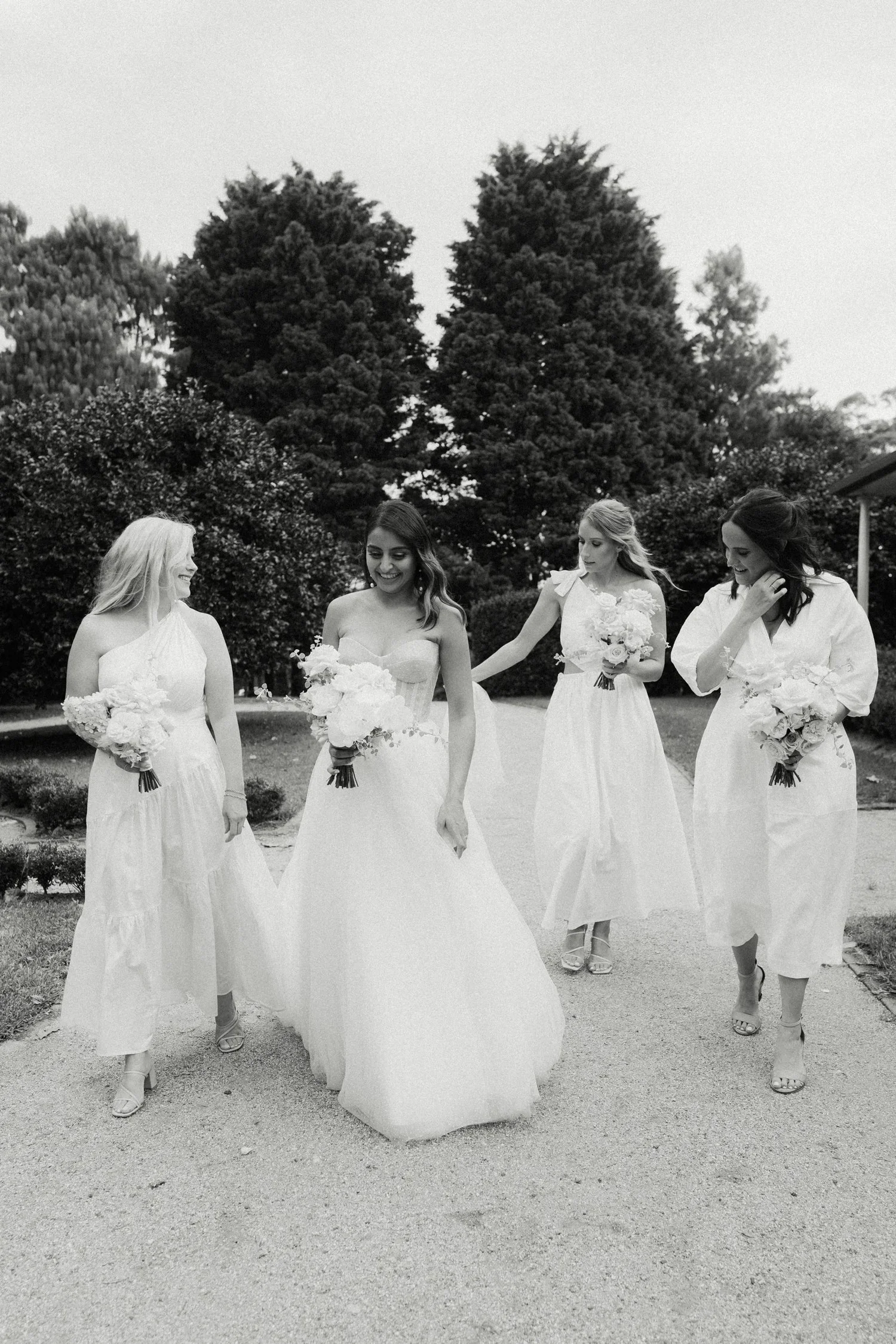 Four women in white dresses and high heels walking outdoors, carrying bouquets of flowers, with trees in the background.