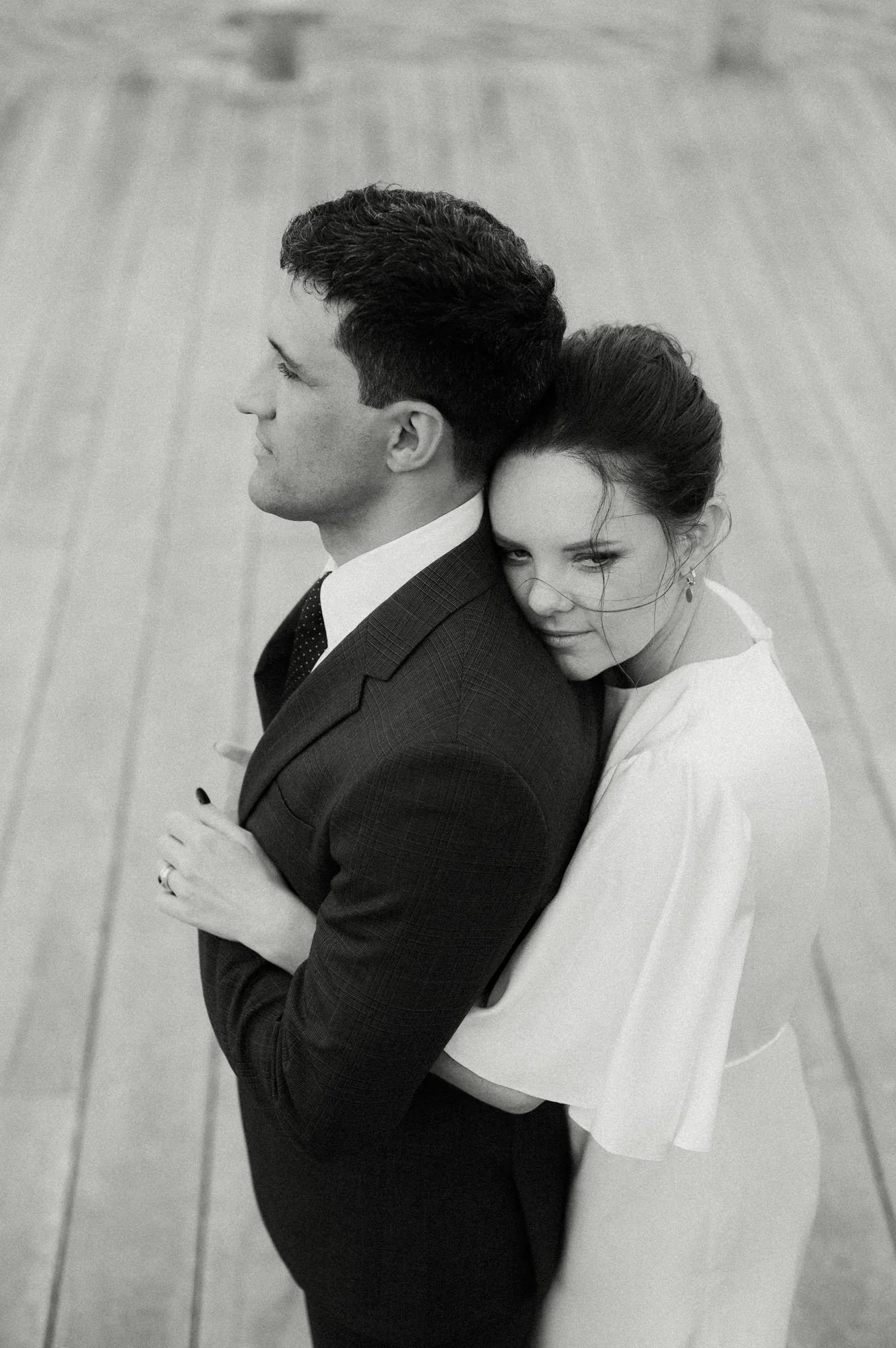 A black and white photo of a couple embracing each other, with the woman resting her head on the man’s shoulder, both with closed eyes, standing on a wooden floor.